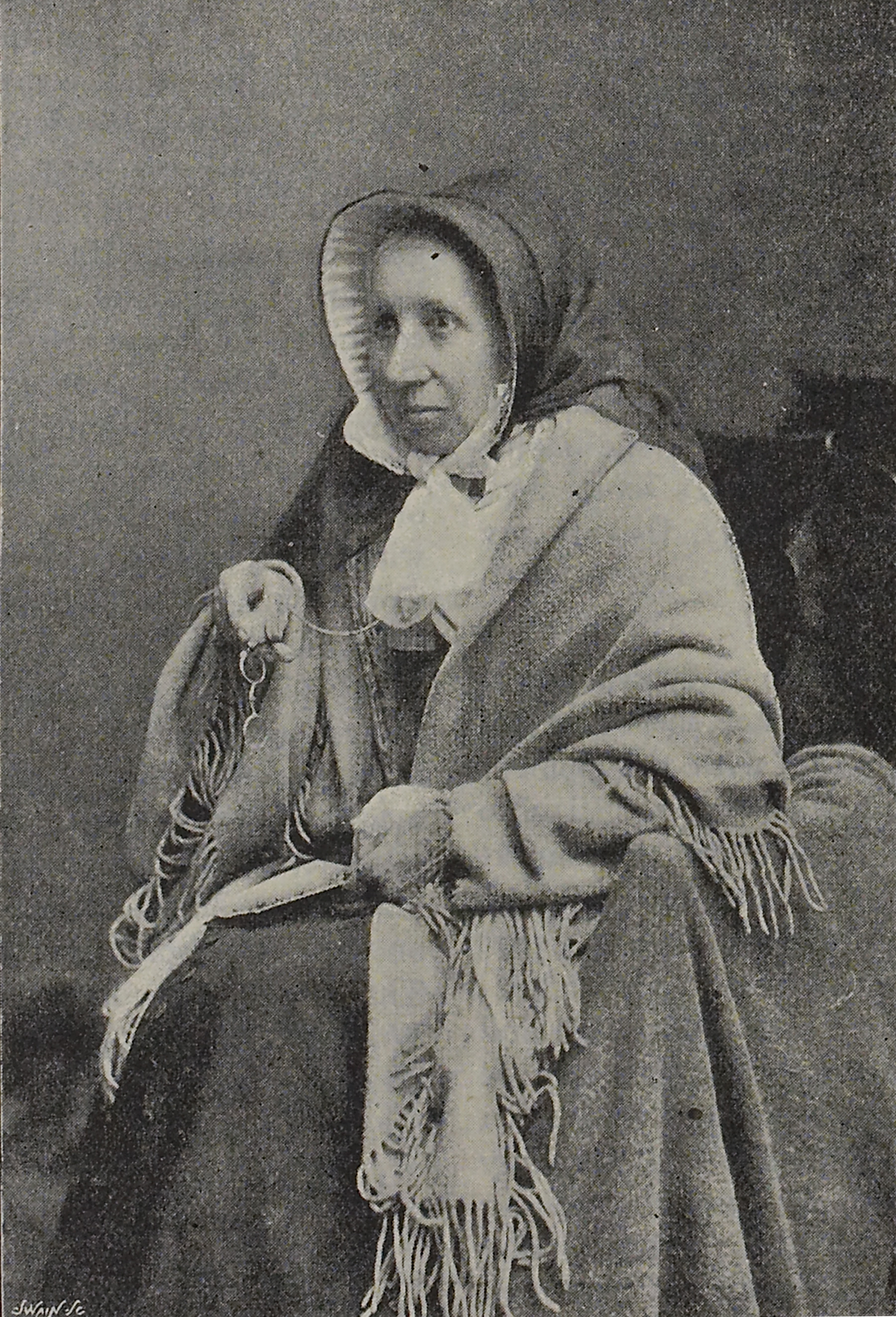 Sepia toned, older newspaper image of woman sat for portrait photo facing at a 45 degree angle. She is wearing period clothing, appears to be wrapped in a shawl and holds her round metal glasses in her right hand.