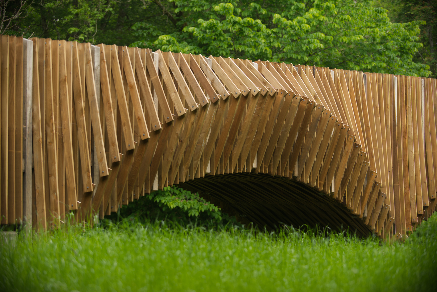 A unique, artistic wooden bridge. The top of the bridge is straight, but there is a distinct arch underneath it, which creates a curve. The bridge is made of individual beams of wood. 