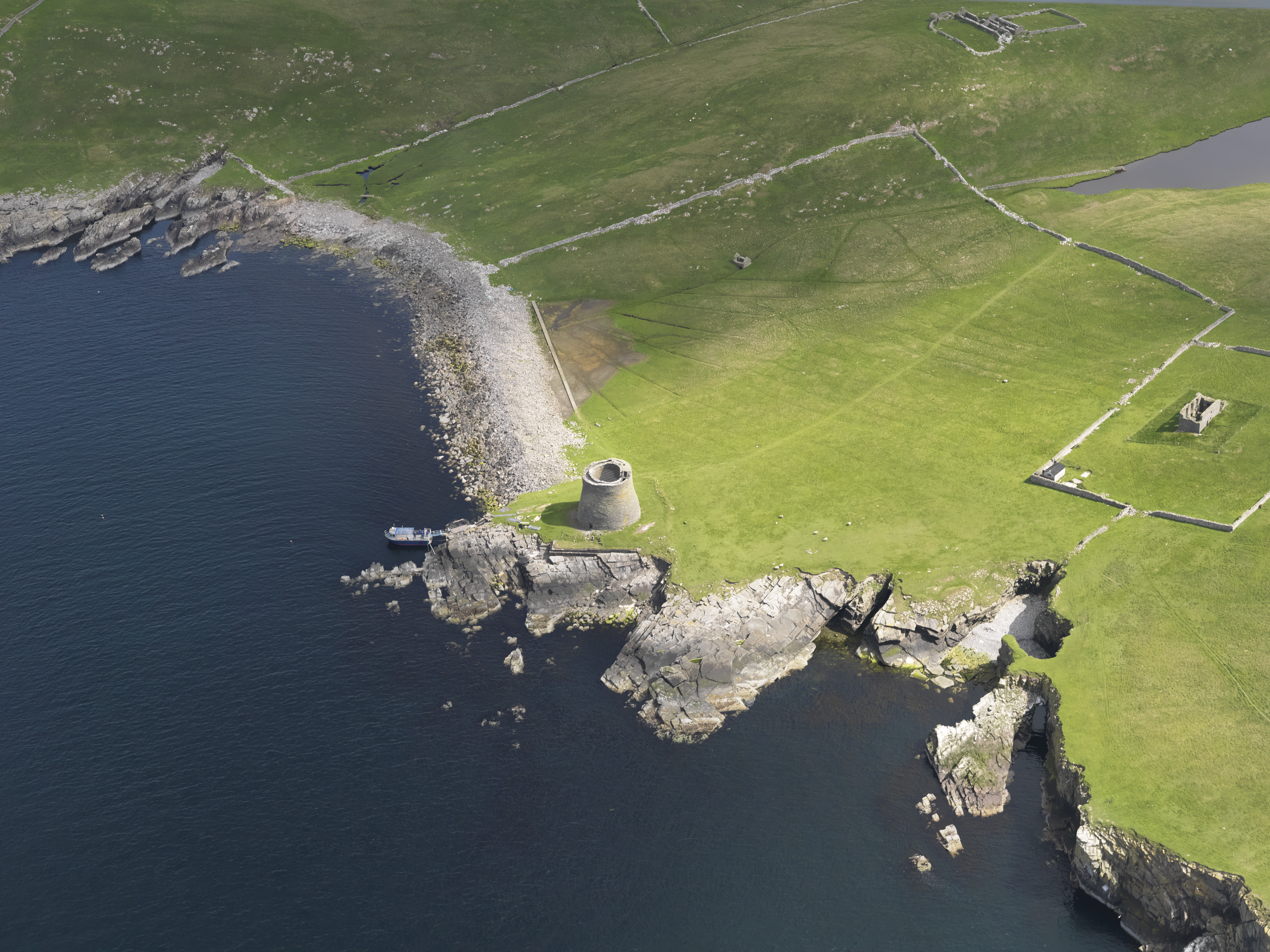 An aerial photograph of a round stone fort stood on a cliff side. To the right and above are green fields with some stone walls running through them. To the left and below are cliff edges and beaches which meet the sea.