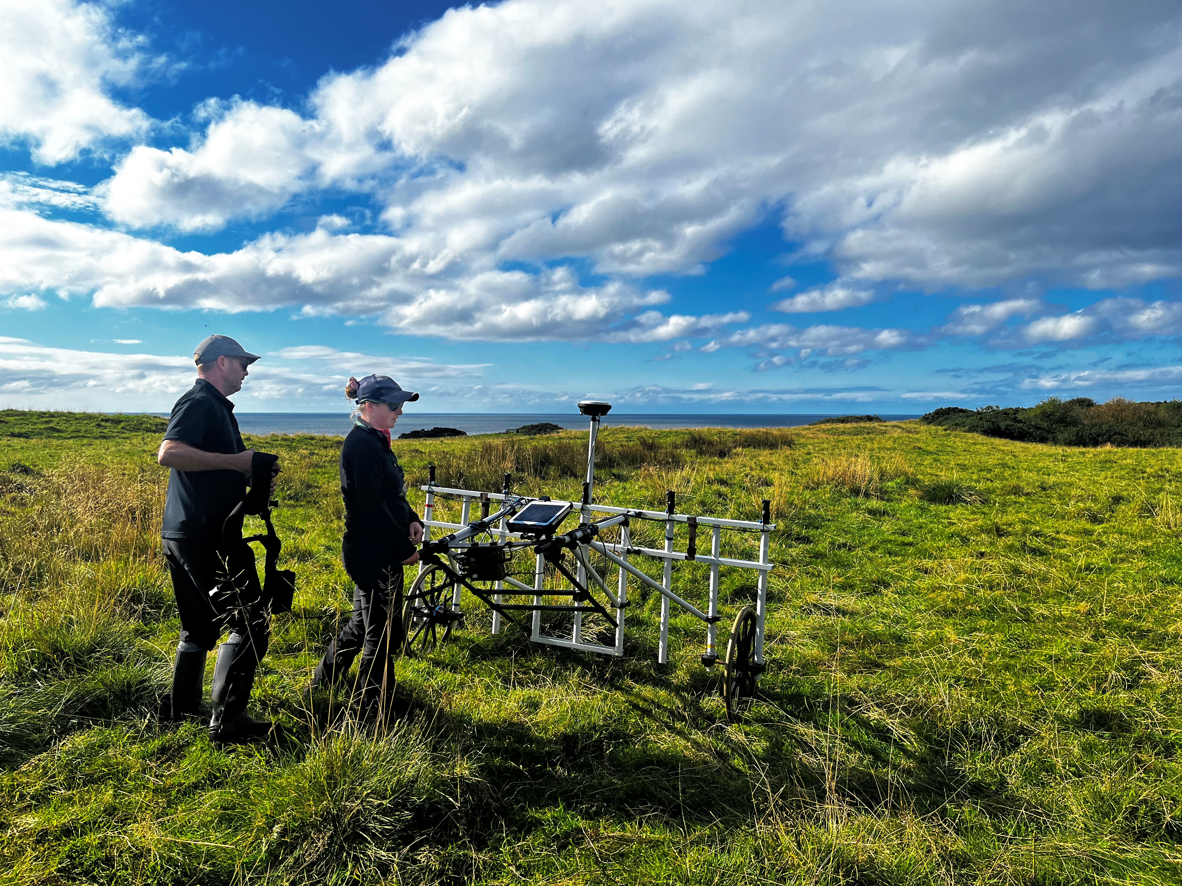 Two field archaeologists stand beside a wheeled geophysical survey frame in a coastal grassland, with the sea and dramatic clouds stretching across the horizon.