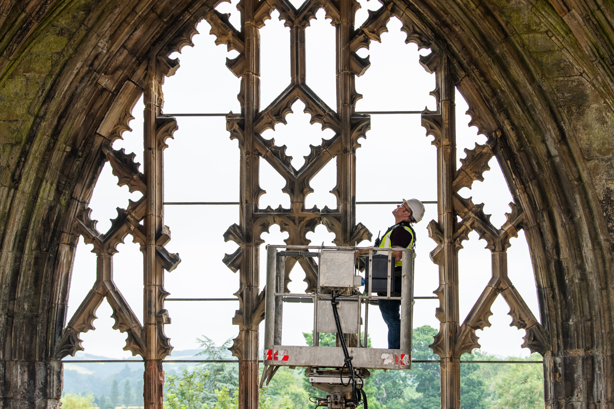 A person wearing personal protective equipment (PPE) while inspecting the stone archway of an abbey from the top of a cherry picker