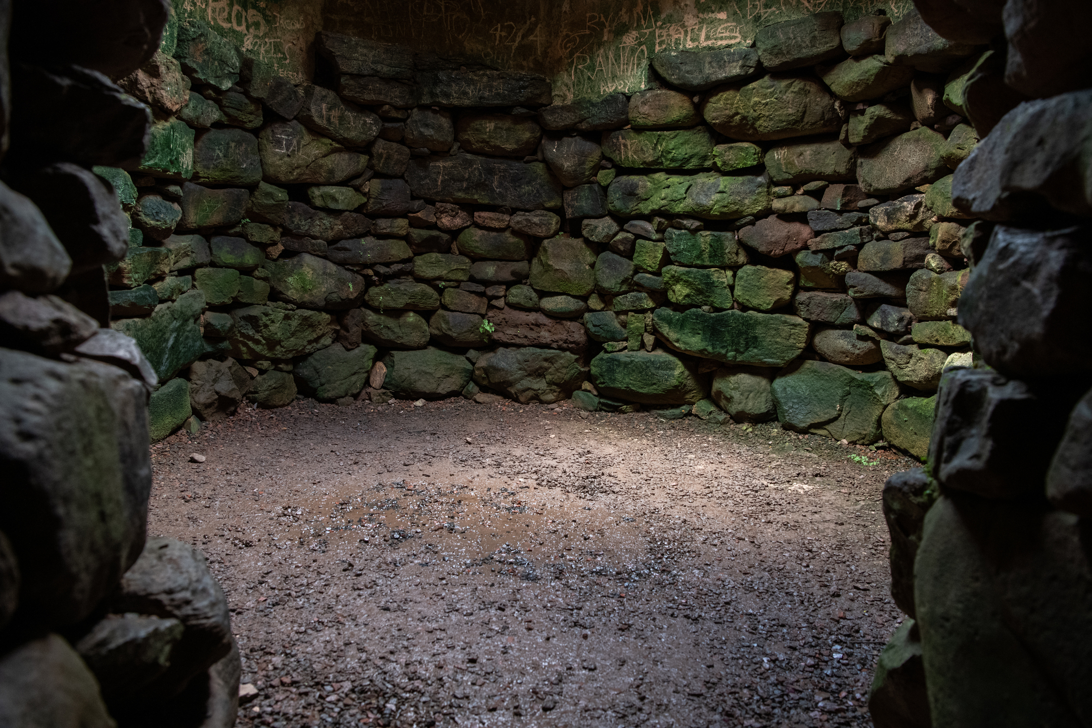 Inside the earth house, showing a small circular stone chamber with rough stacked walls and a dirt floor, dimly lit from the entrance.