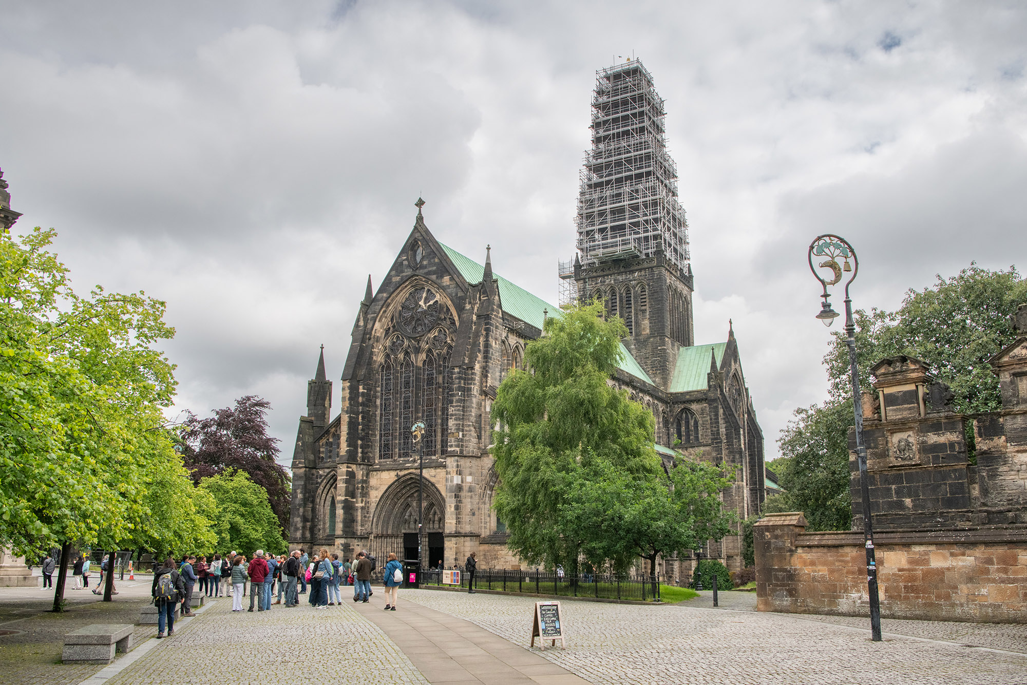 Glasgow Cathedral viewed from the cathedral precinct, with visitors gathered near the main entrance on a cobbled square. The Gothic stone building features large stained-glass windows and a central tower enclosed in scaffolding as conservation work takes place. Mature trees frame the scene, while ornate street lamps and historic stone walls surround the cathedral beneath a cloudy sky.