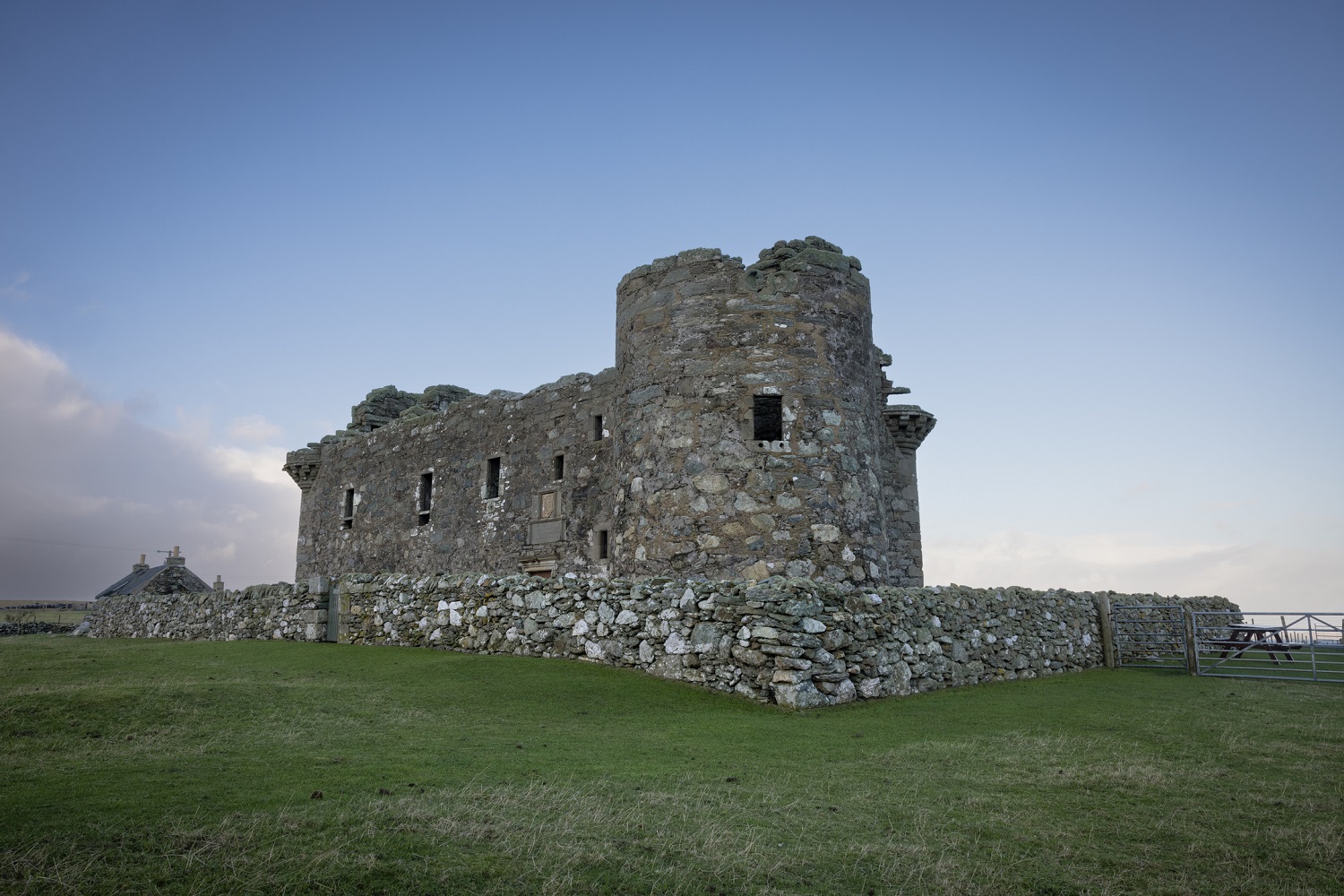 Exterior view of Muness Castle, a historic stone structure with rounded towers and thick, weathered walls. The castle sits on a grassy hill enclosed by a low stone wall. 