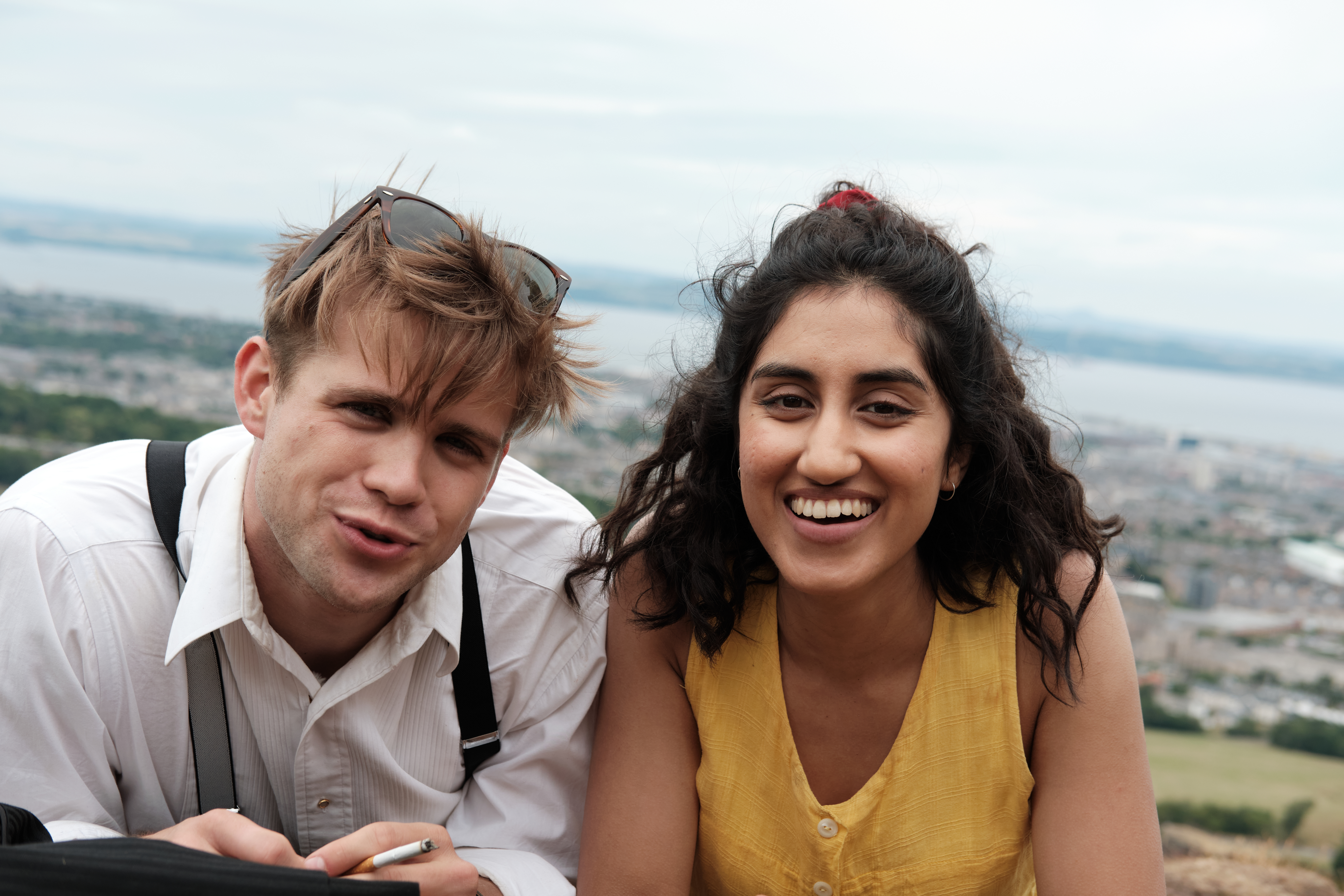 Two young adults sitting outdoors on a hill with a city and water in the background. They smile toward the camera; the man has messy hair and sunglasses on his head, and the woman wears a yellow sleeveless top.