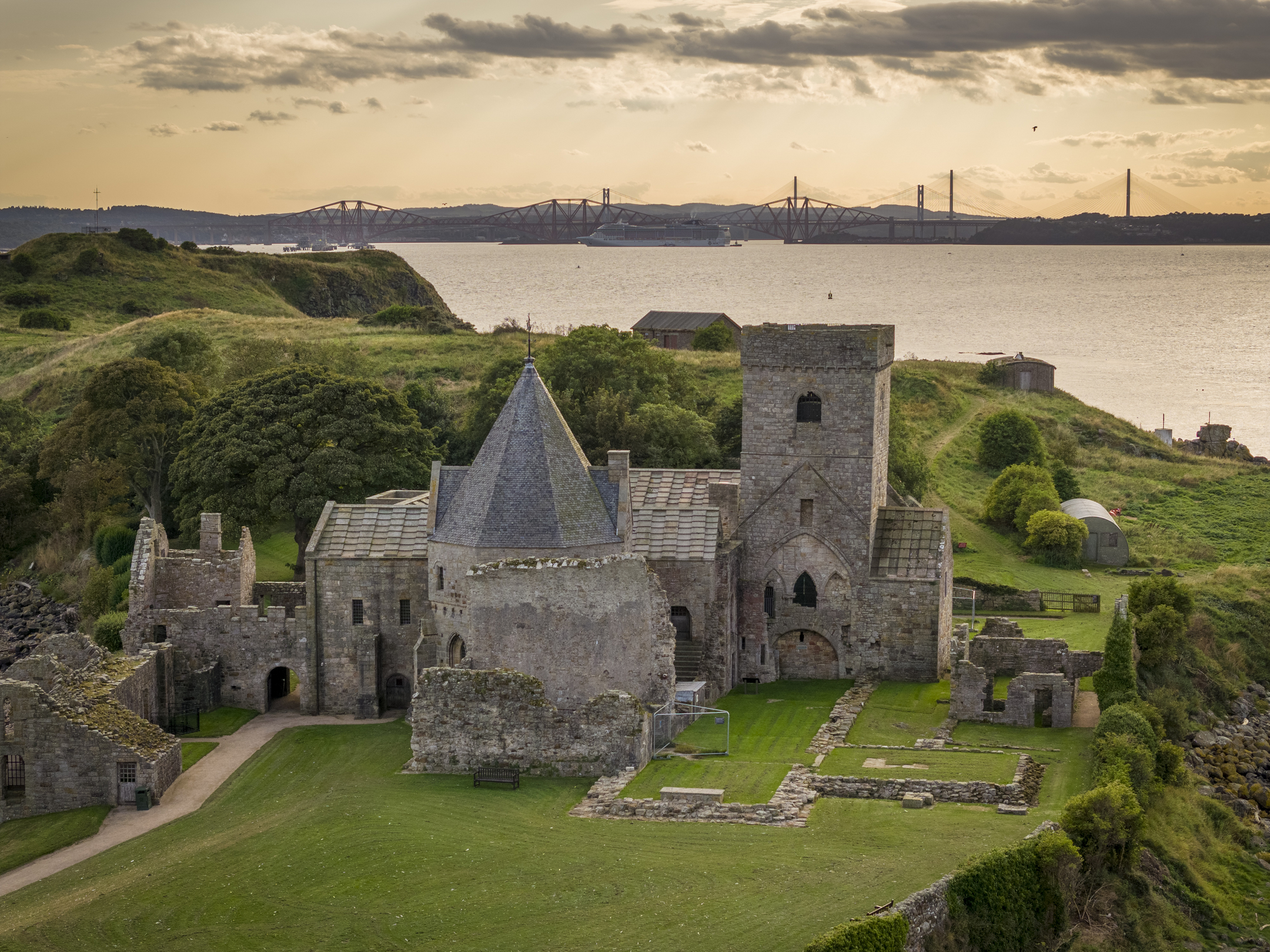A large, historic abbey with some ruin areas and some slate roofs, on the edge of the water with the forth and clackmannanshire bridges in the background