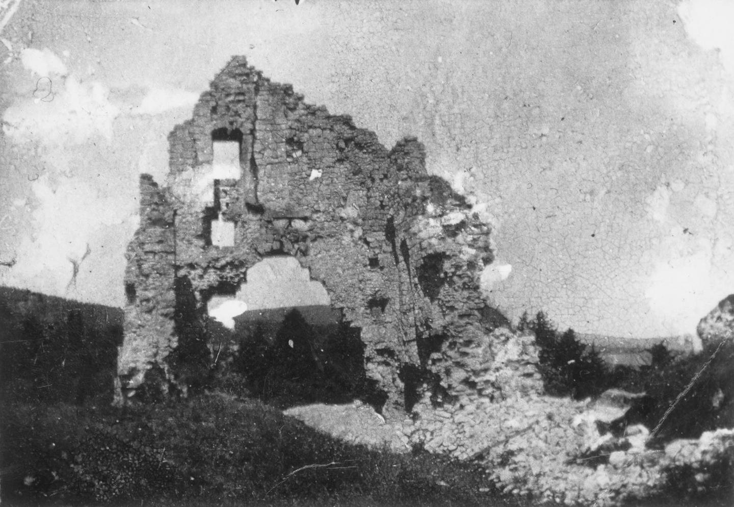 A black‑and‑white photograph showing a heavily collapsed stone wall of Kildrummy Castle. A large arched opening remains in the centre, with broken masonry scattered around. The wall stands alone on a grassy slope.