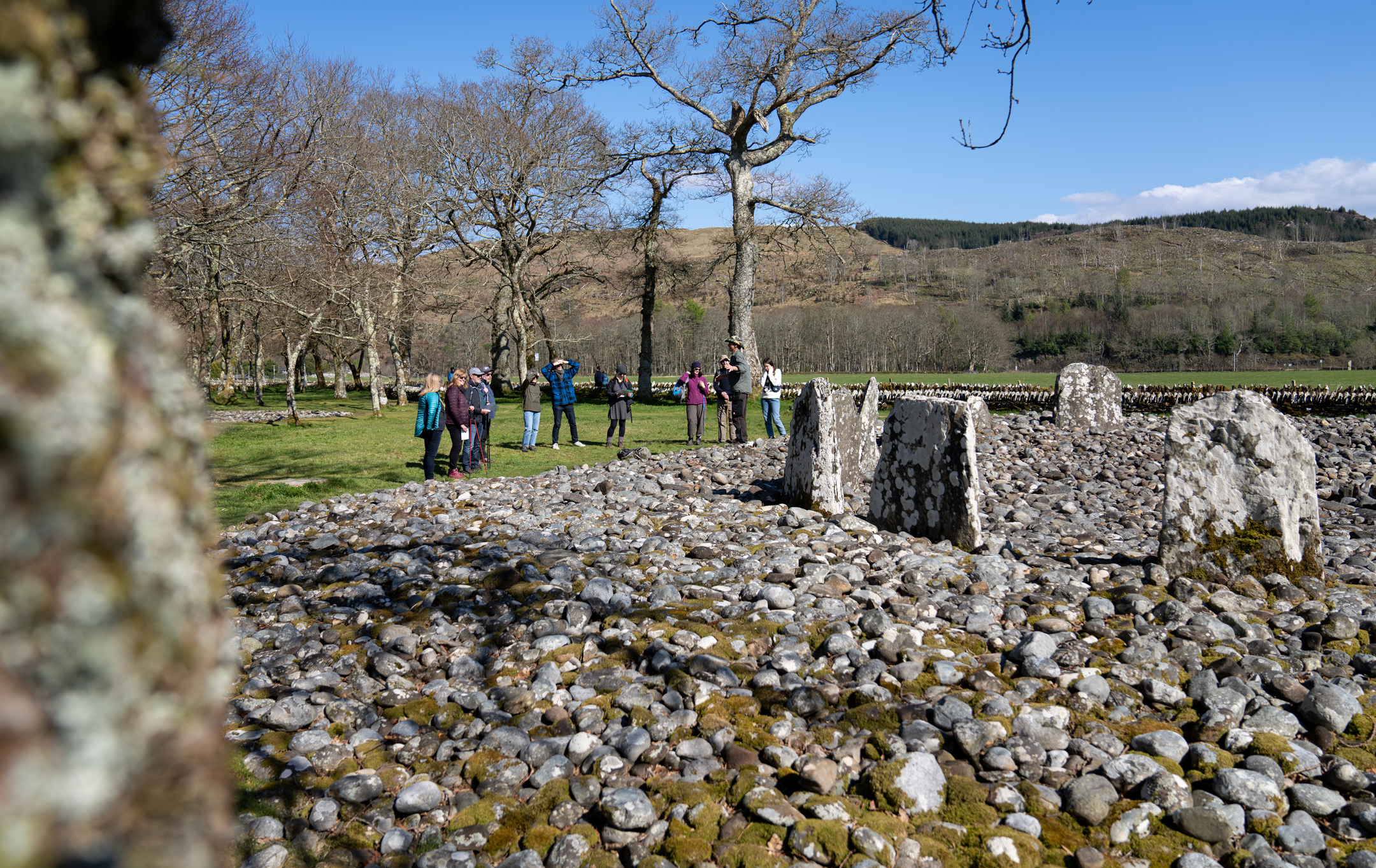 Area covered in stones, with standing stones in it, and in the distance a crowd of people, around 10 of them, looking at the stones