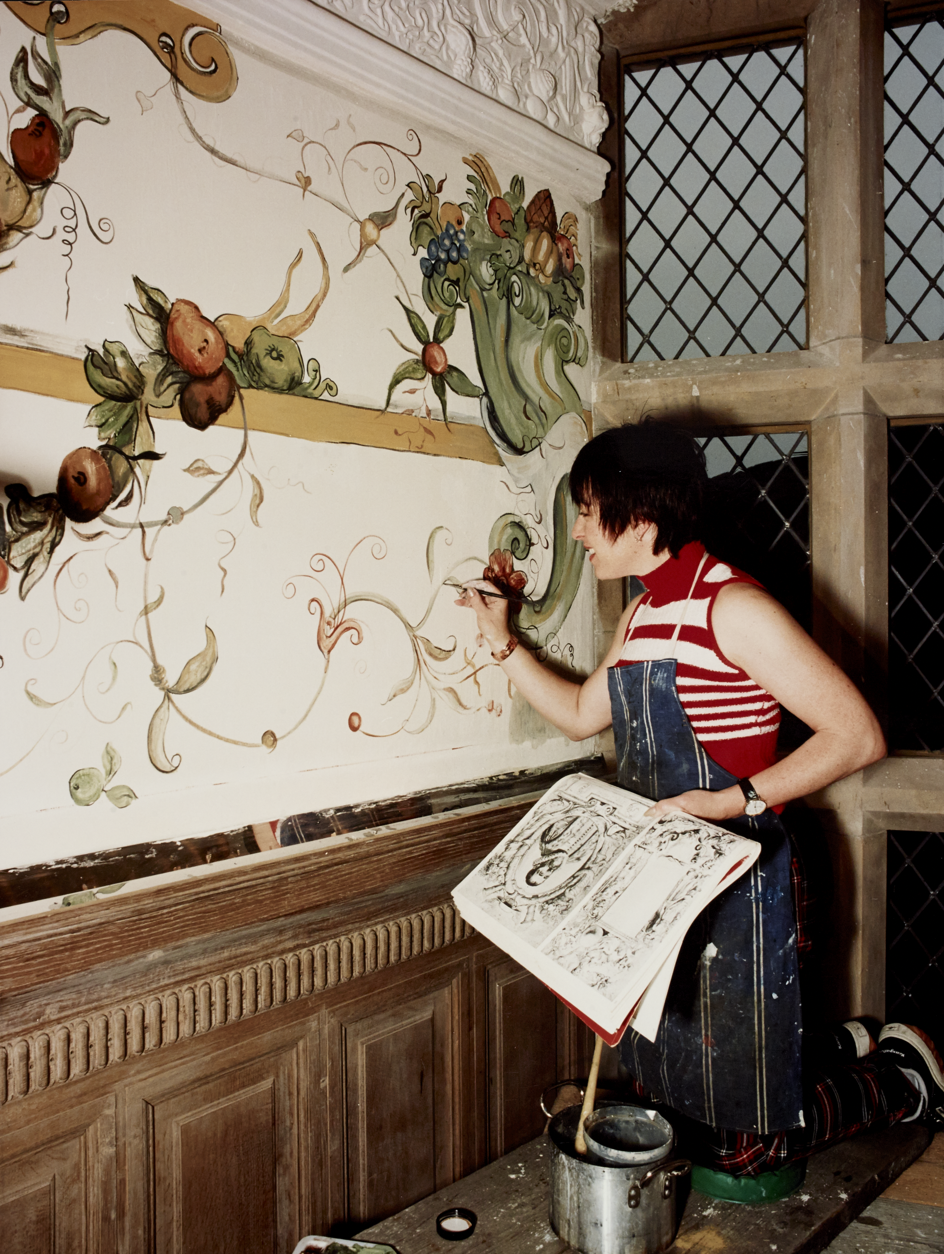 Woman paints wall based on reference image she is holding. Wall decoration features plants, flowers and fruit