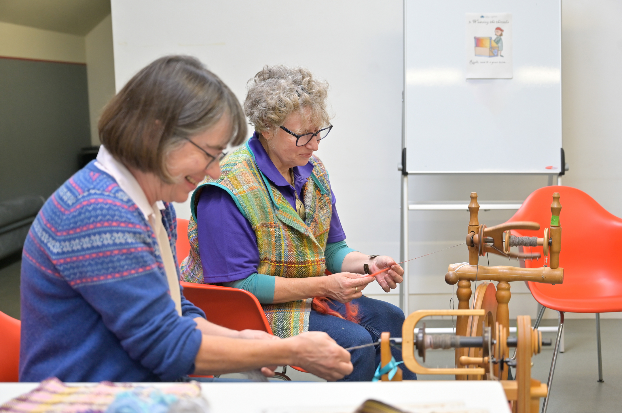 Two people sit working with wool at a wooden spinning wheel, focusing on preparing fibres in a bright room