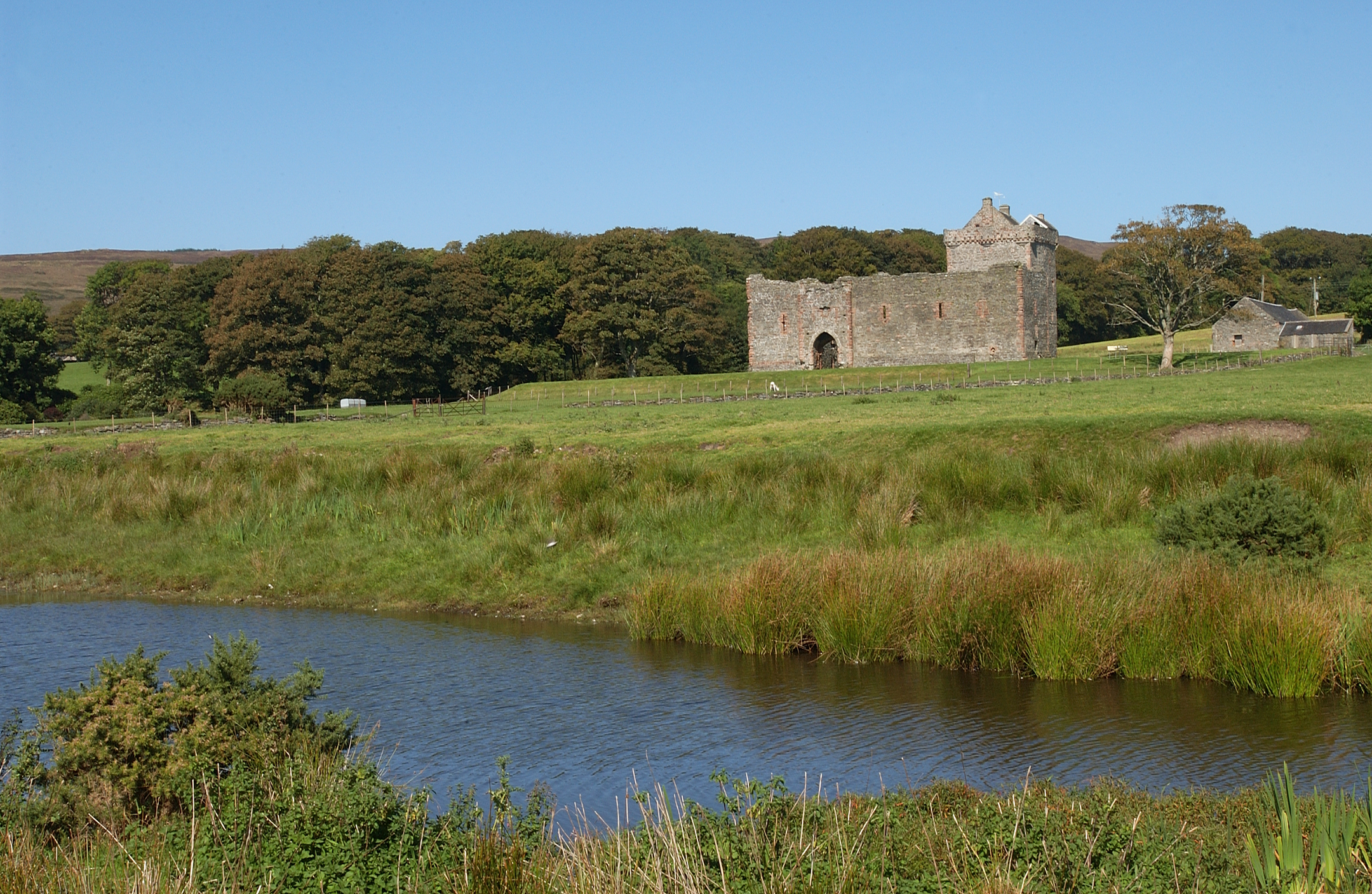 Distant view of a stone castle ruin sitting in a field, photographed across a river or pond with trees behind the castle.