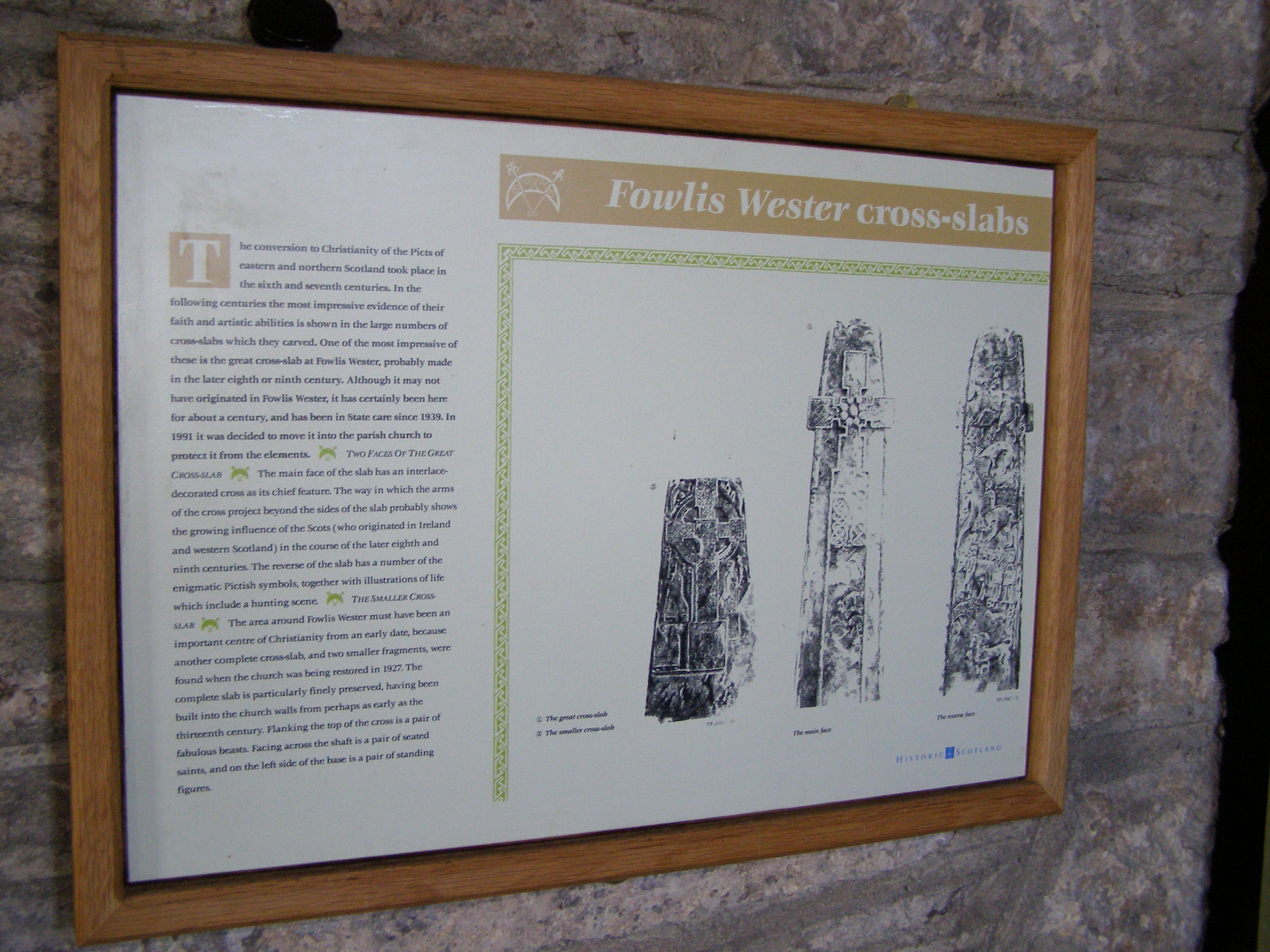Interpretive panel titled ‘Fowlis Wester cross-slabs’ mounted in a wooden frame on a stone wall. The sign includes descriptive text about the historical significance of the stones and three black-and-white illustrations of cross-slabs with intricate carvings. The text explains their origin and artistic details.