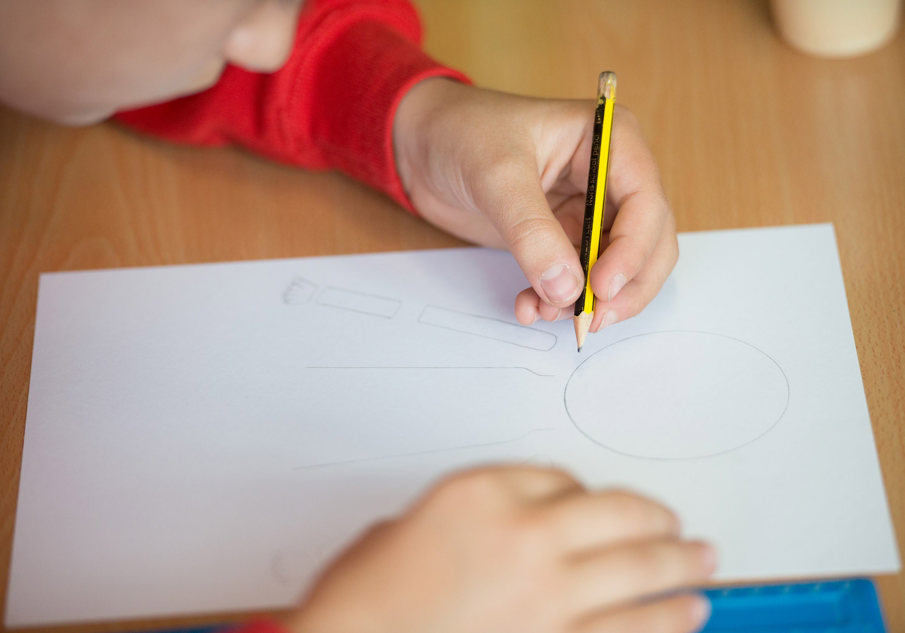 A child wearing a red jumper sitting at a table drawing on a piece of white A4 paper