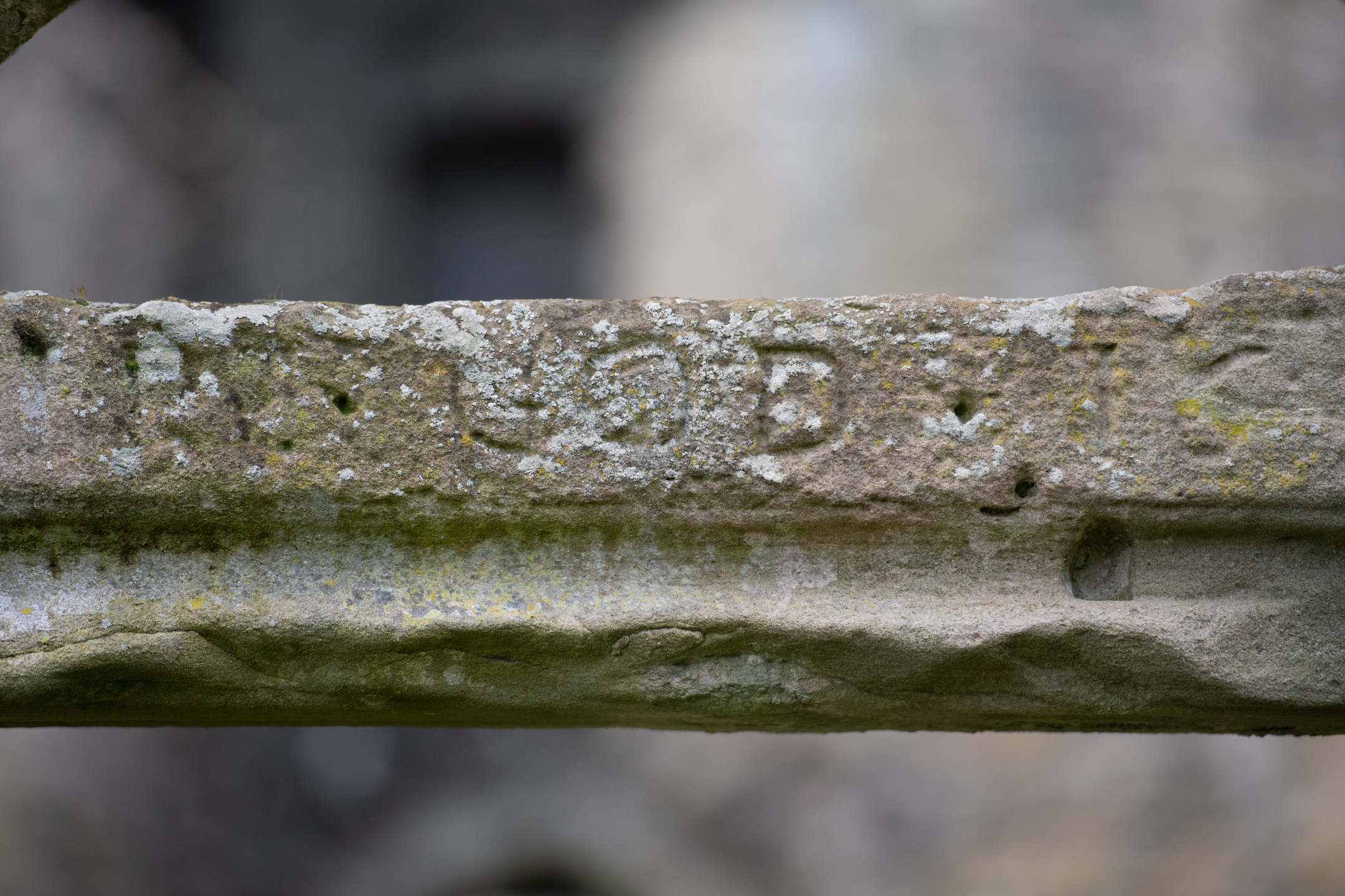 Detailed close-up of a carved inscription on a stone lintel. The letters appear worn and partially illegible, with moss and lichen growing on the surface. 