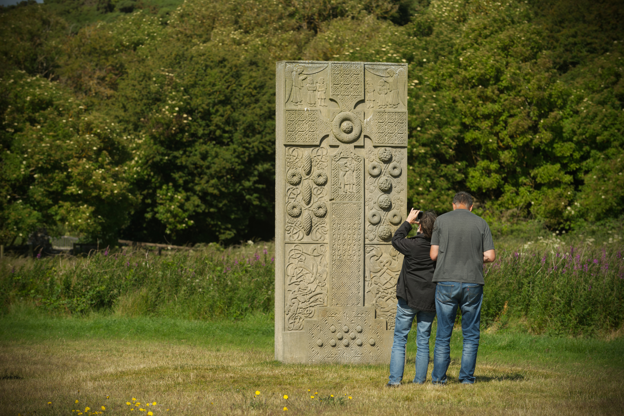 Two visitors standing close to the Hilton of Cadboll replica stone, examining and photographing its detailed carvings. The stone is set in a grassy area with wildflowers and dense green foliage in the background.