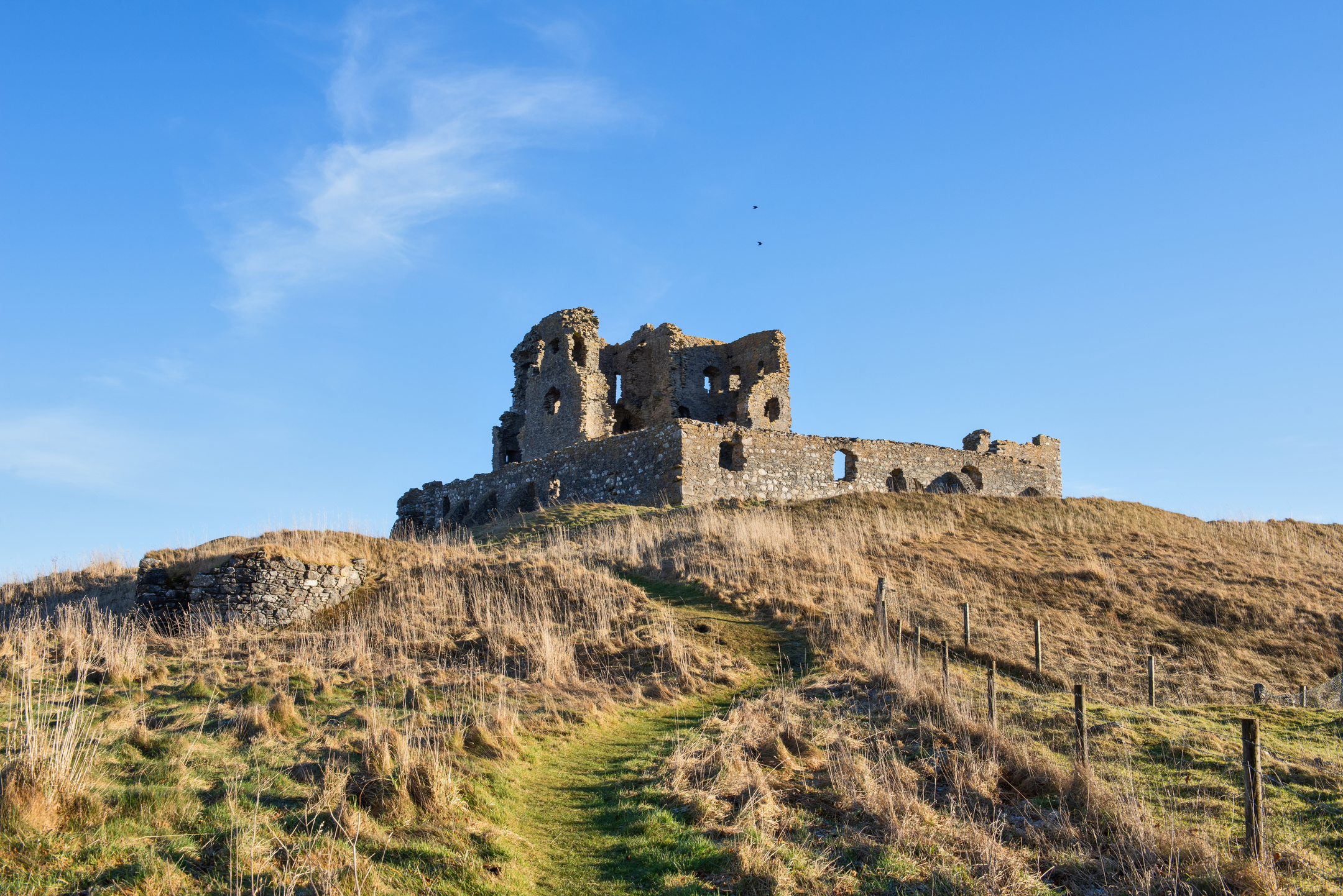 A narrow and winding grassy path leads across a hillside to a ruined castele