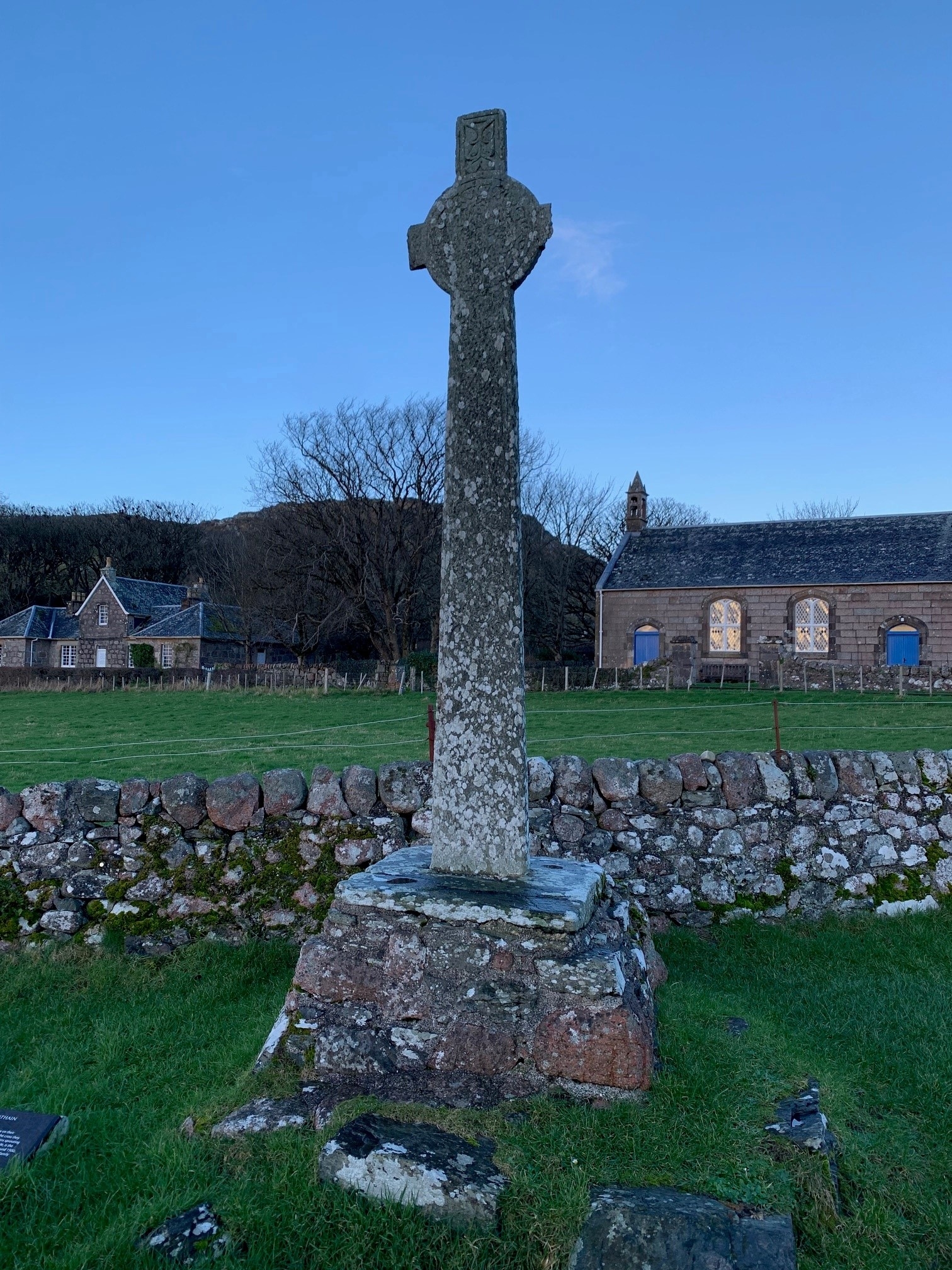 Front view of a tall stone cross with a circular ring at the intersection of the arms, rising from a multi‑layered stone base. A dry‑stone wall runs behind it, with a field, bare trees, and stone buildings visible in the background beneath a clear blue sky.