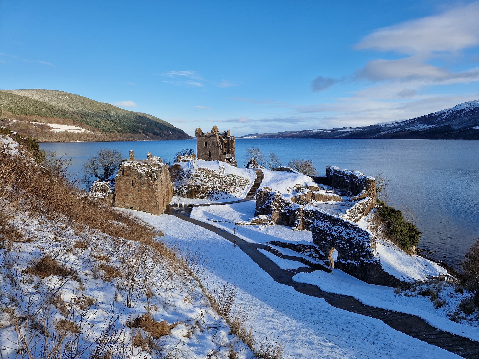Blue skies over the ruins of a castle covered in snow. It is dramatically located on the shores of a large loch. 