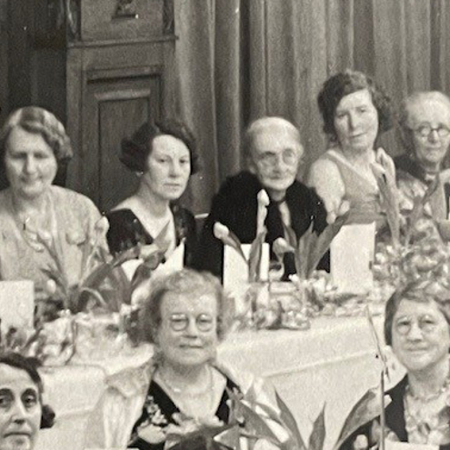 Groups of women at some kind of reception sit at tables decorated with flowers. There is a row of five women of varying ages in the rear of photo, along a straight table. 