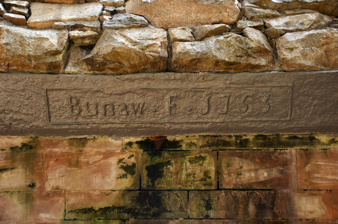 Close‑up of an old stone wall with a rectangular panel carved with the inscription ‘Bunaw F. J. 1563’. The carved section sits beneath rough stones and above weathered bricks stained with moss