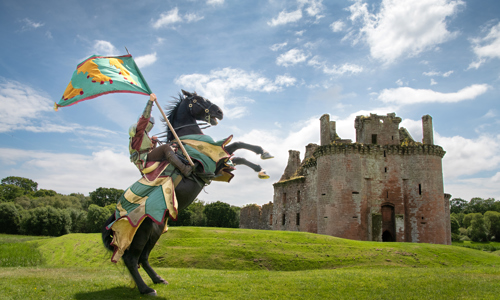 A medieval knight in armour sits atop a black horse that is rearing up its front legs whilst stood on a grassy green field. The knight holds a green and yellow flag in his right hand. ruinous castle is visible in the background, its a sunny day and the sky above is blue with some white clouds.