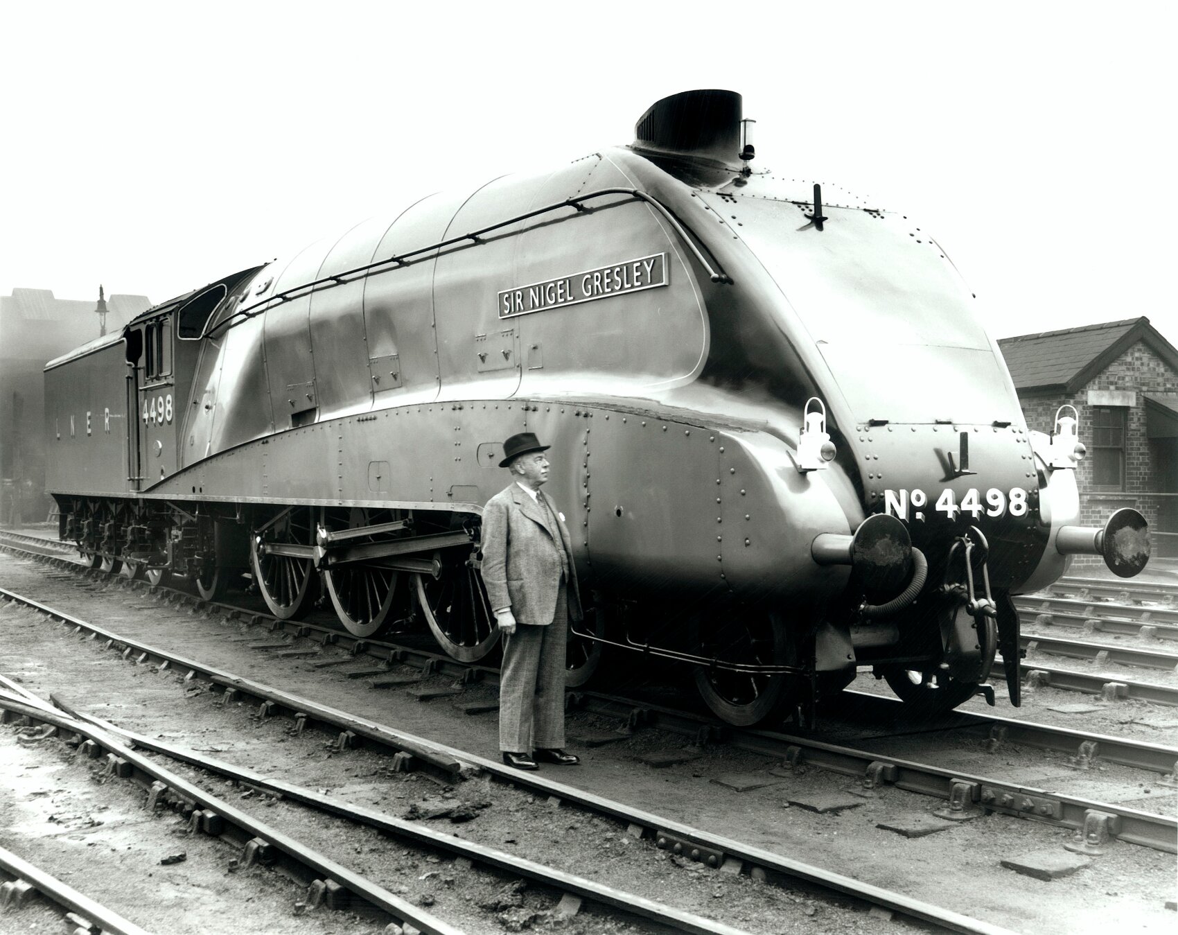 Black and white photograph of a person in a grey suit and dark trilby hat standing next to, and looking up at, a steam locomotive engine in a goods yard.