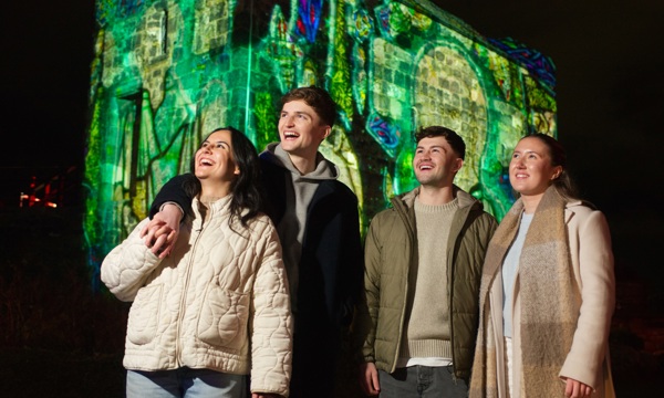 A group of four young people are standing looking up at the distance and smiling, wearing jackets, jumpers and one person is wearing a scarf. In the background is a castle lit up in green with images projected onto the walls. 