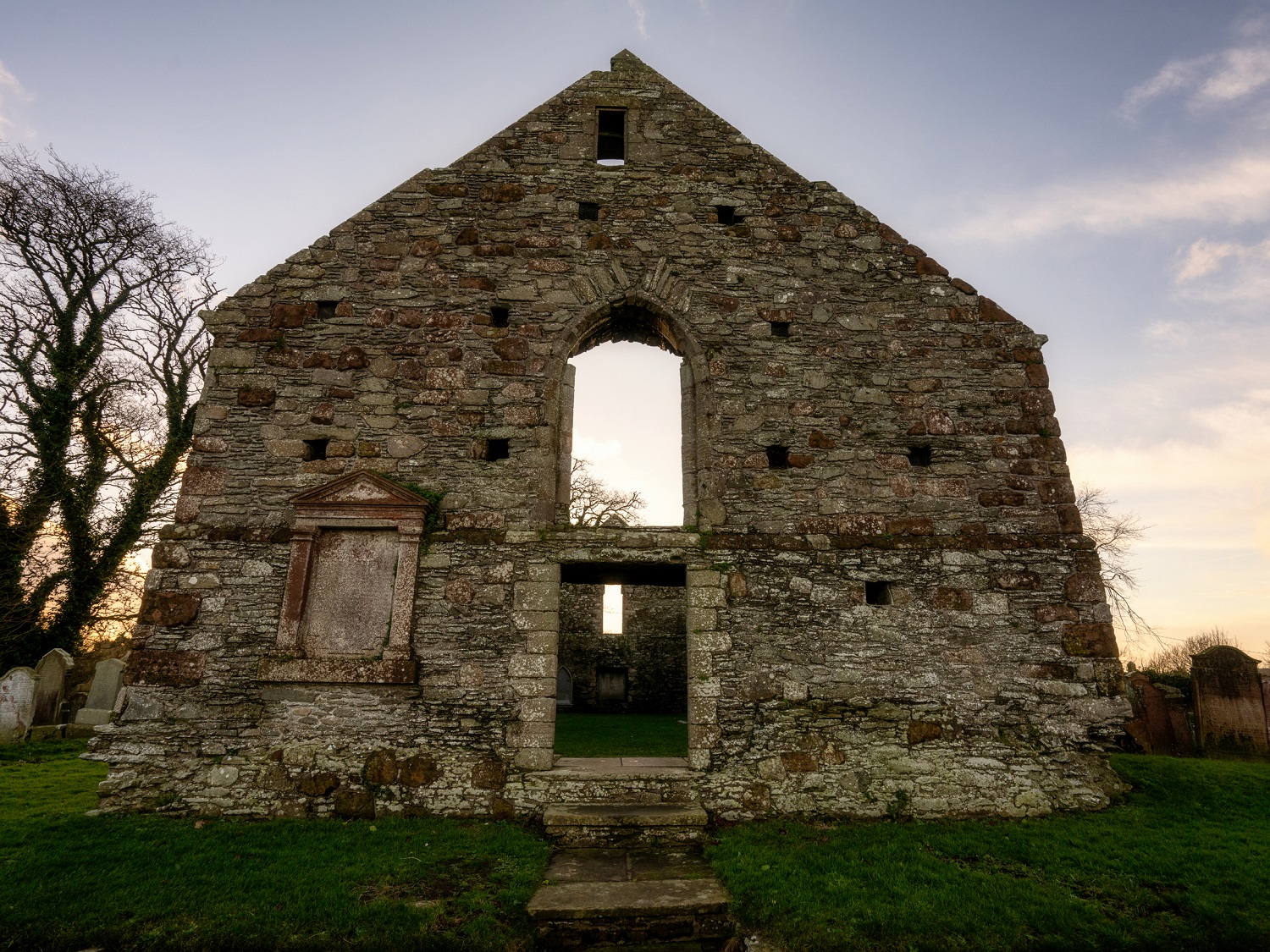 Front view of Whithorn Priory’s gabled stone façade, with a large arched opening and smaller doorway below, surrounded by grass and gravestones.