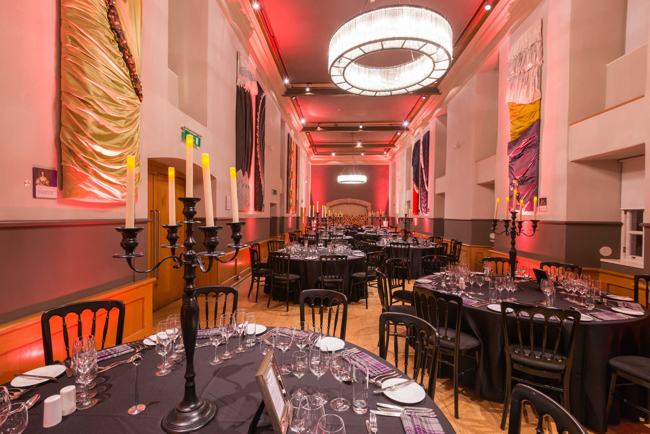 Many circular tables are set ready for dinner guests in a long rectangular room. 