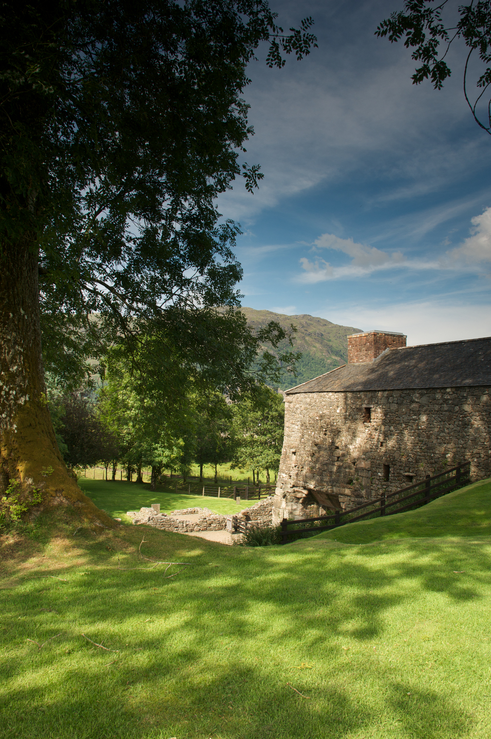 an old stone building with small windows and a chimney on top, sitting on a grassy slope next to a large tree