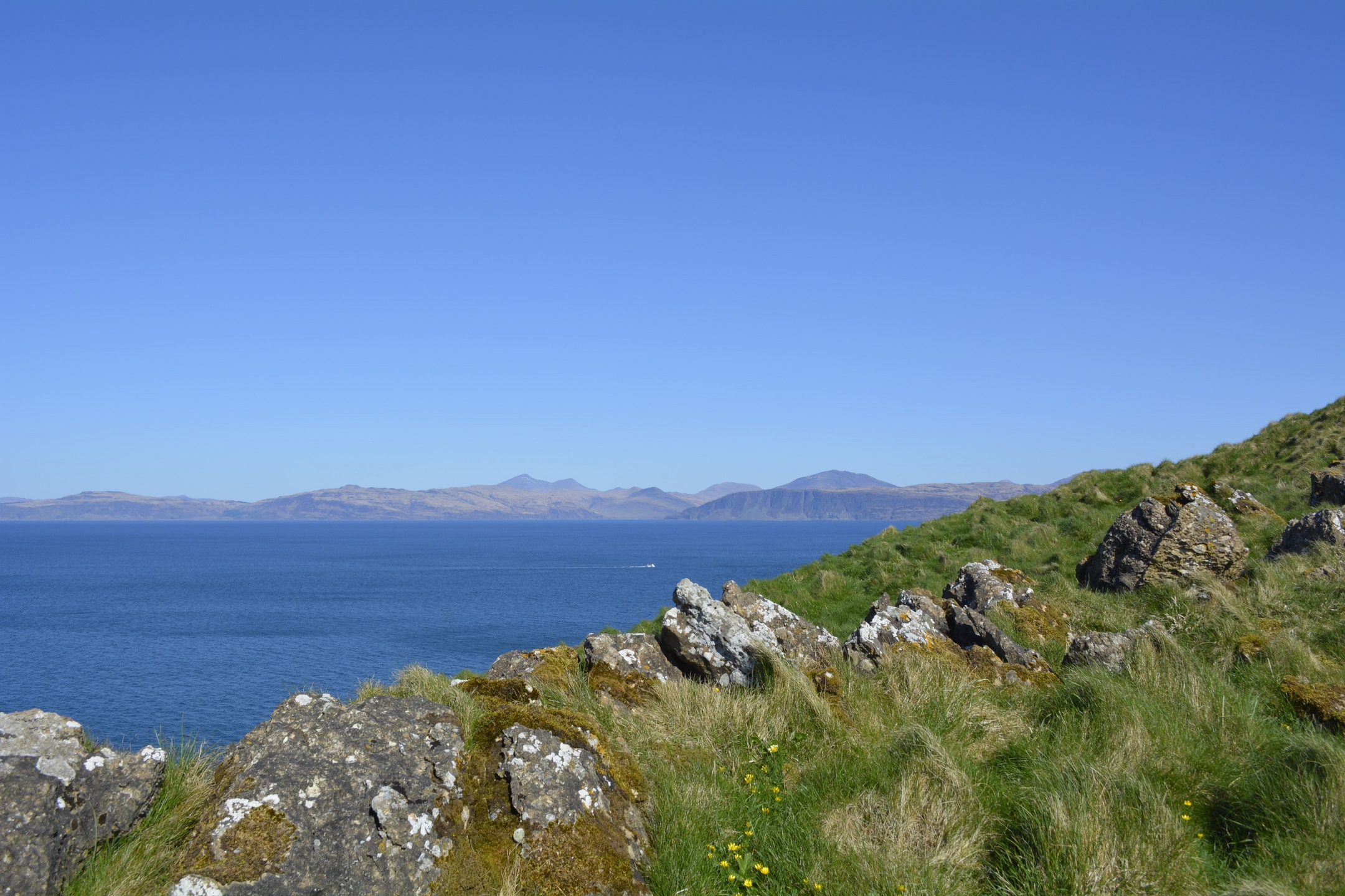 Panoramic view of a calm blue sea with distant mountains under a clear sky, seen from a grassy, rocky coastline.