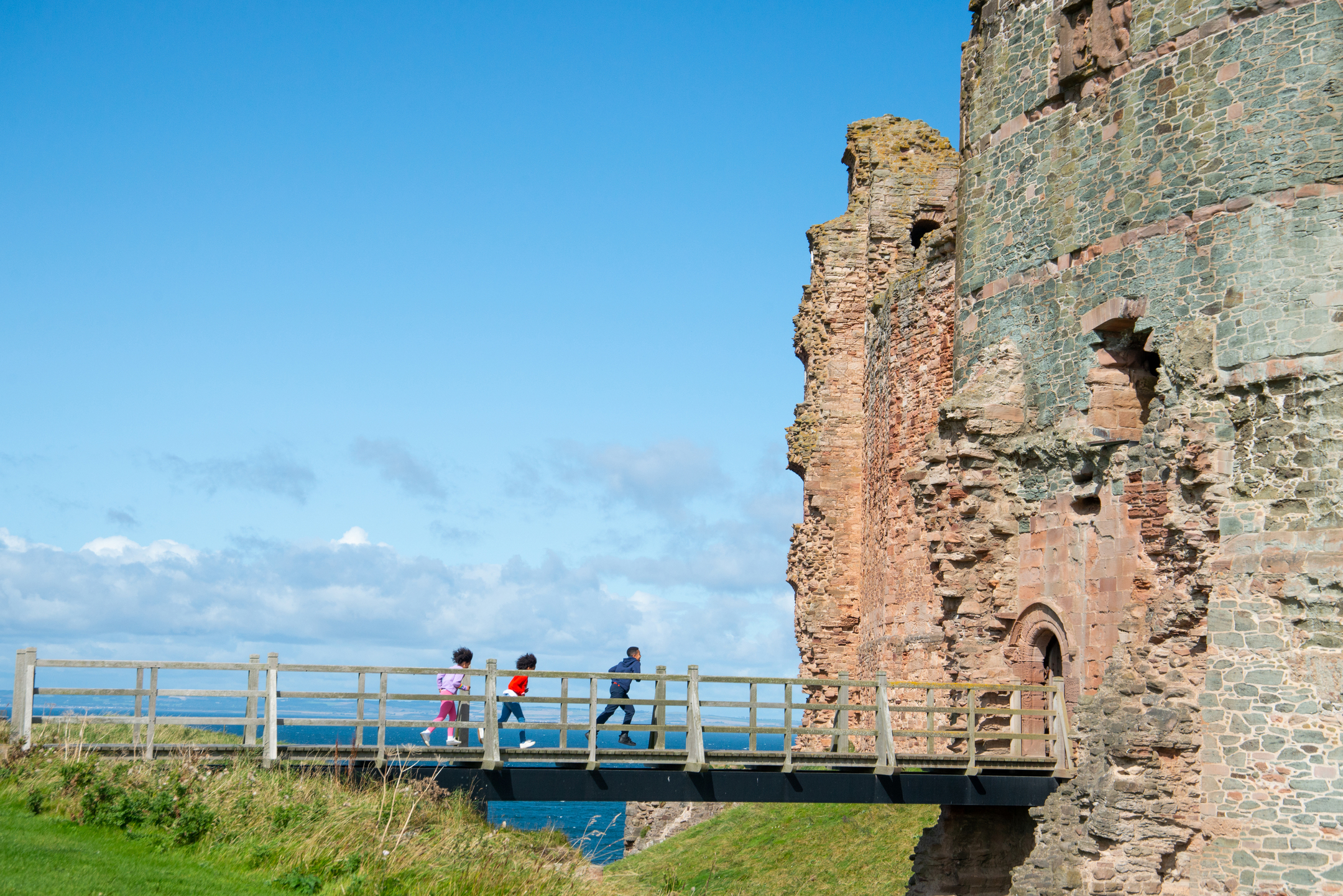 Three children cross a bridge from a grassy area towards a castle entrance