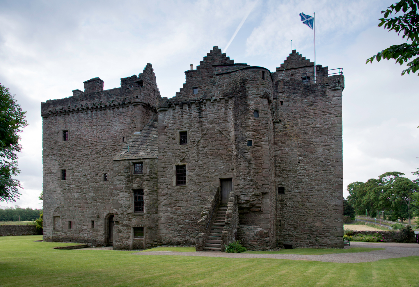 Exterior of a medium size castle. There are two doors, one with a staircase leading up to it. There are multiple windows. It sits on a grass landscape, with a saltire flag flying.