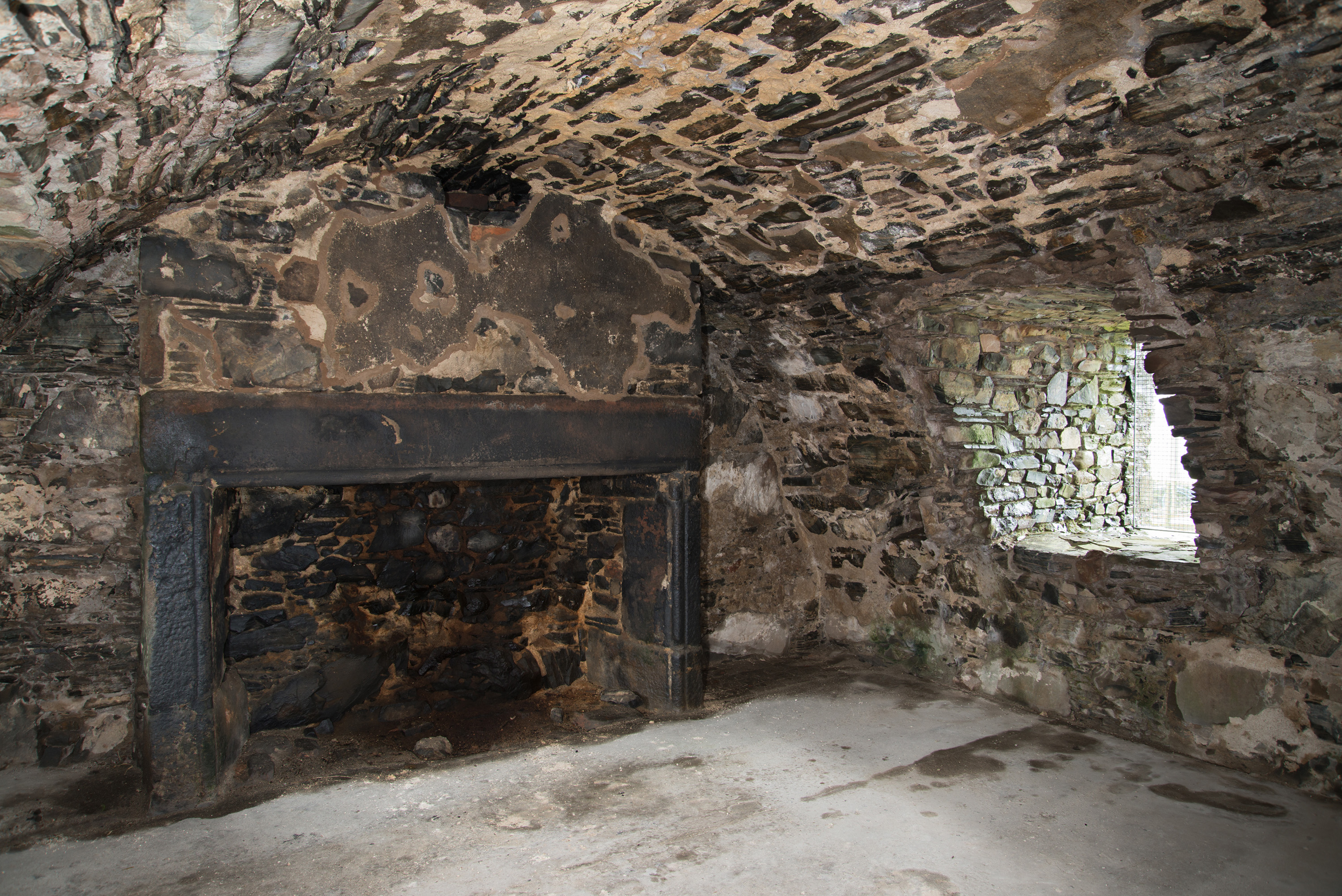 Interior view of a stone room in Drumcoltran Tower, featuring a large, soot‑blackened fireplace set into a thick stone wall. The fireplace has a heavy, dark lintel supported by two upright stone jambs, with charred stonework visible inside the hearth. The walls and vaulted ceiling are built of irregular stones, mottled with age and moisture. A small, deep-set window on the right lets in natural light, illuminating the rough textures of the room.