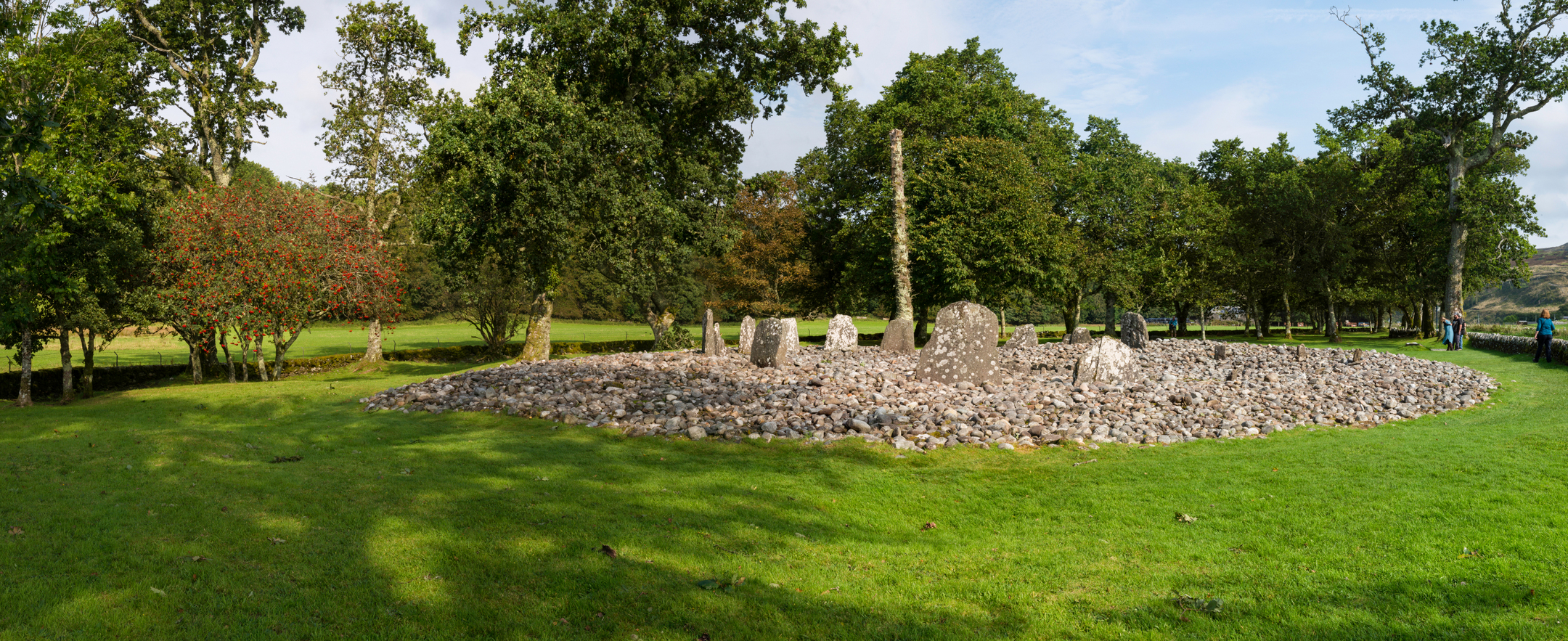 A panoramic view of Temple Wood Stone Circle set in a grassy clearing. The ring of stones is surrounded by a wide spread of pebbles, with tall trees forming a dense backdrop. A few visitors stand on the right side of the image.