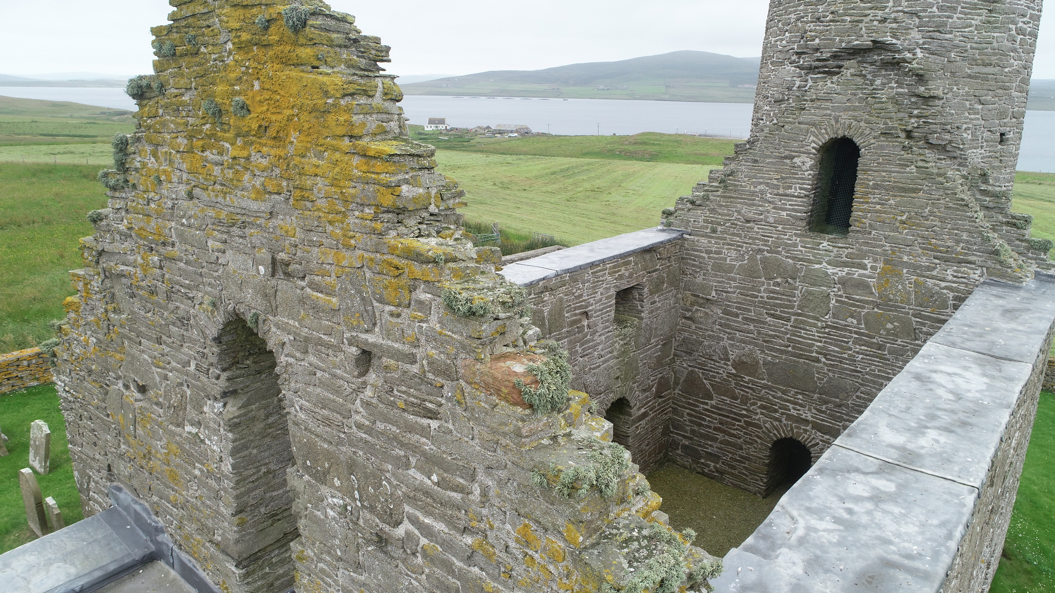 Elevated view of the church ruin showing stone gable walls and the round tower, with fields and water behind