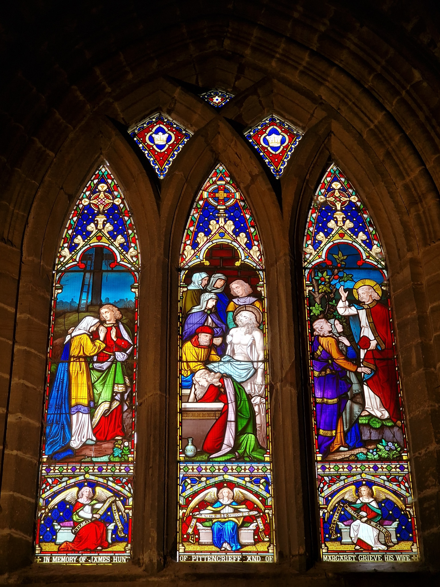A vibrant three‑panel stained‑glass window depicting biblical figures in rich red, blue, and gold tones, framed by dark stone.