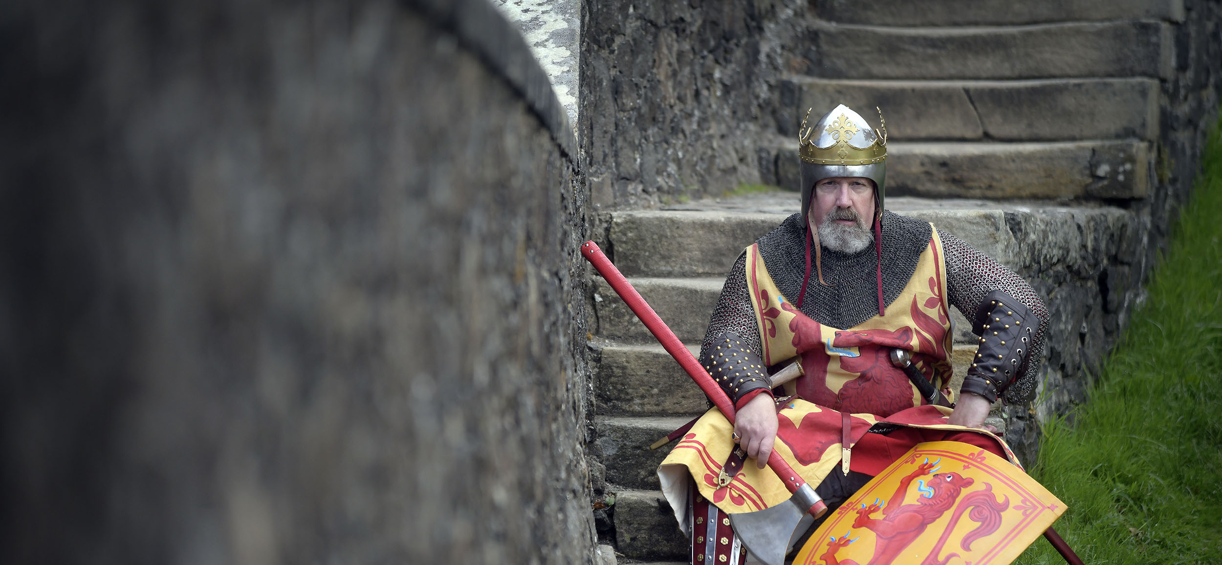 A costumed performer in chain mail with helmet and shield, looking stern sitting on concrete steps