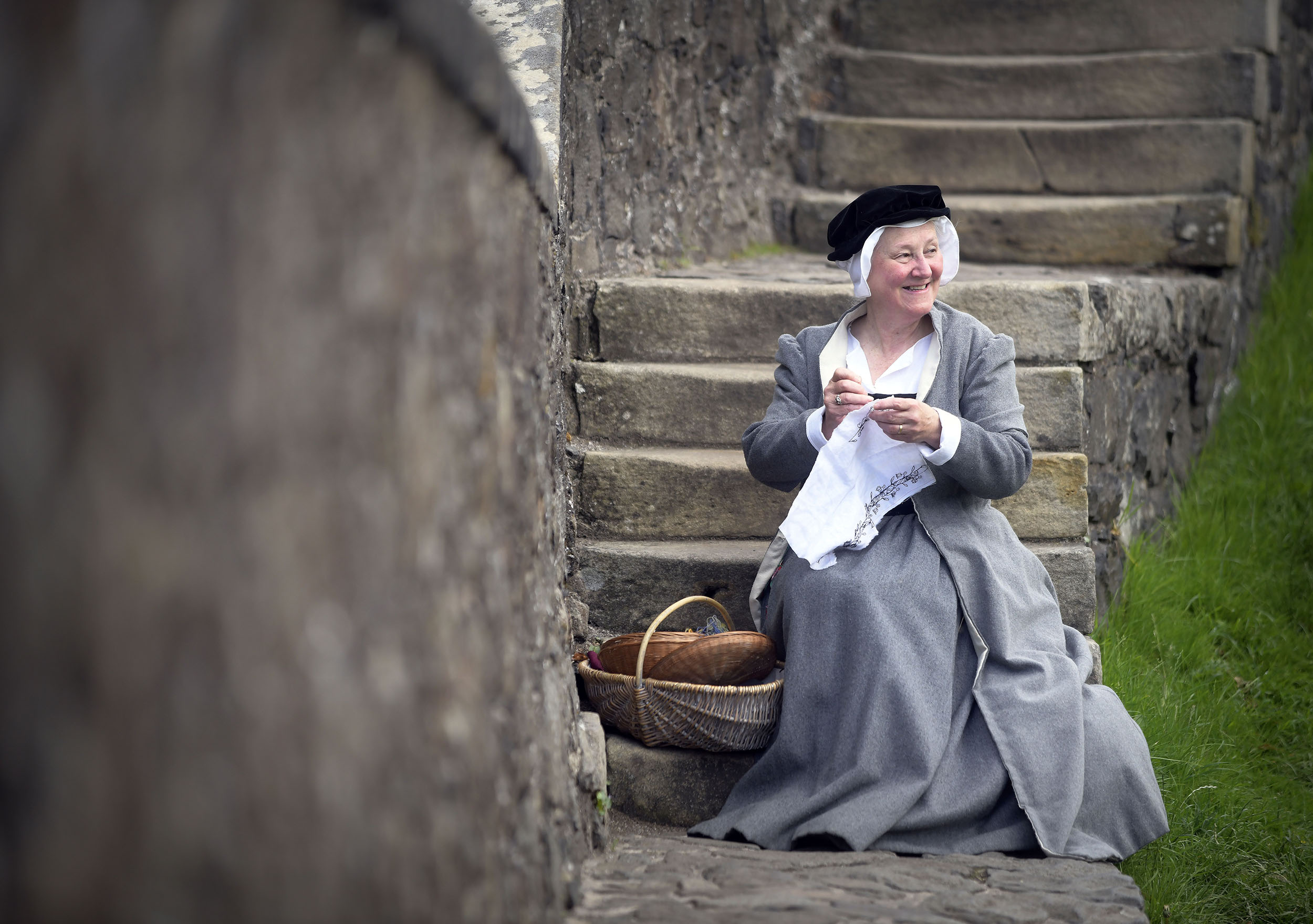 A costumed performer as a maid sitting on large concrete stairs  with some wicker baskets and stitching a piece of cloth