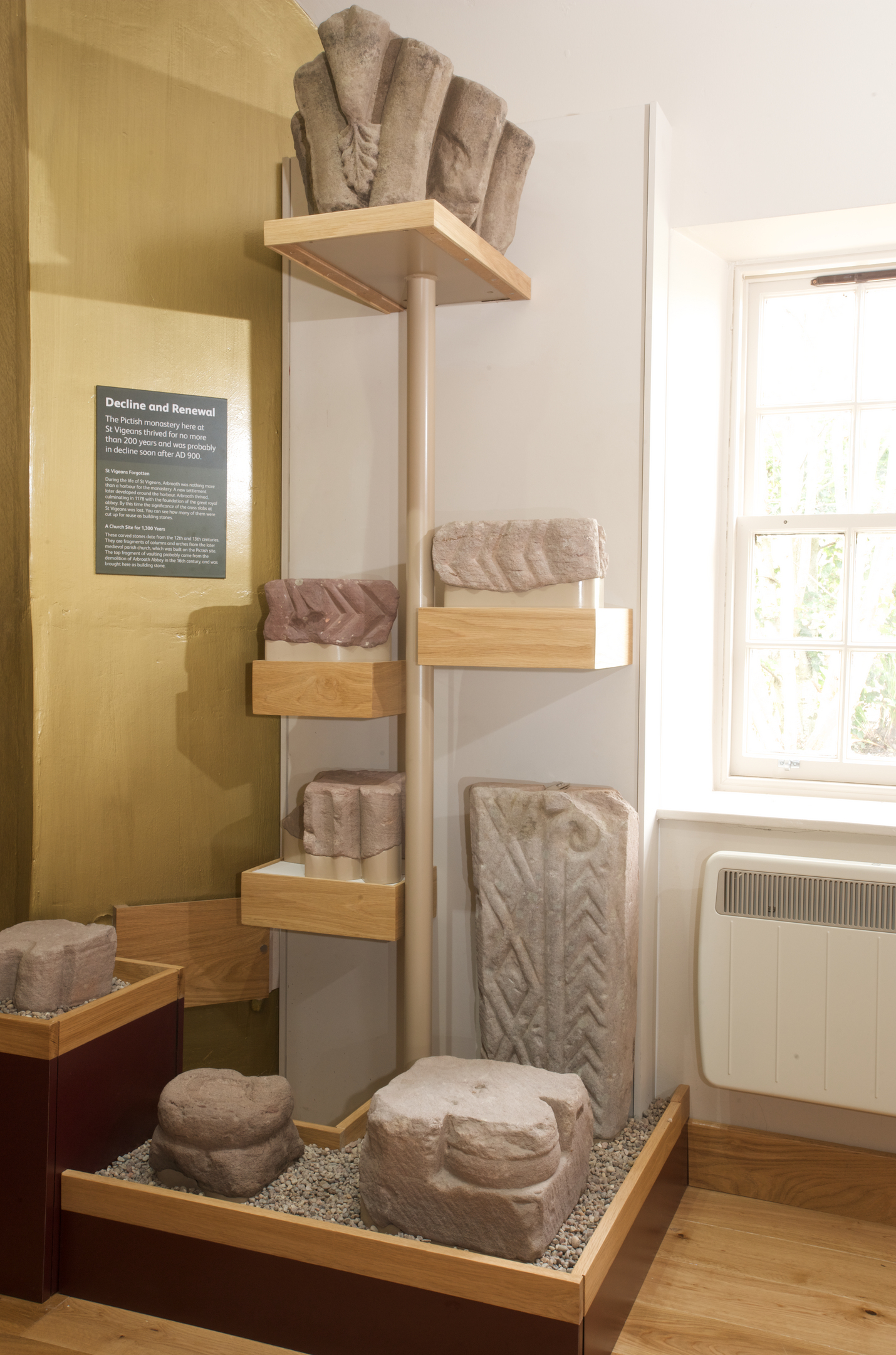 Collection of carved Pictish stone fragments displayed on wooden shelves and stands, including decorated blocks and a tall slab with zig‑zag patterns. The exhibit label titled “Decline and Renewal” is mounted on the wall behind.