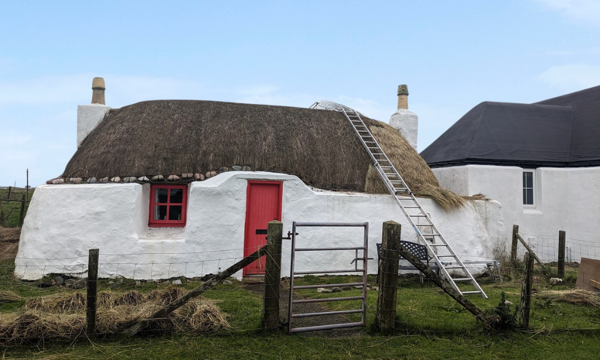 A small stone built, thatched, lime washed cottage with a red front door. A ladder leans on the roof and it looks like the cottage is half way through being re-thatched.