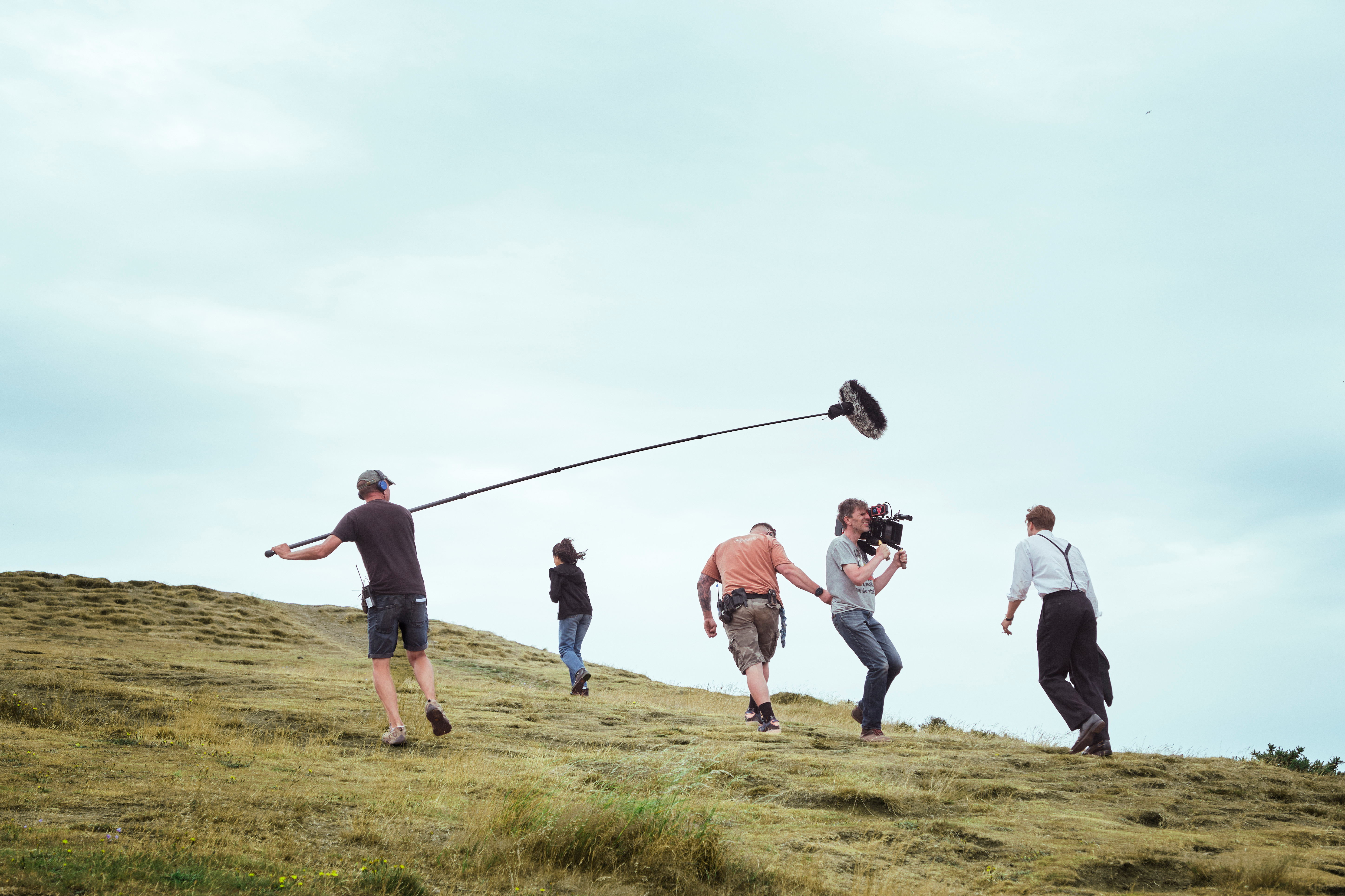 Five people including a film crew and an actor film a scene will walking up a grassy hill under an overcast sky.