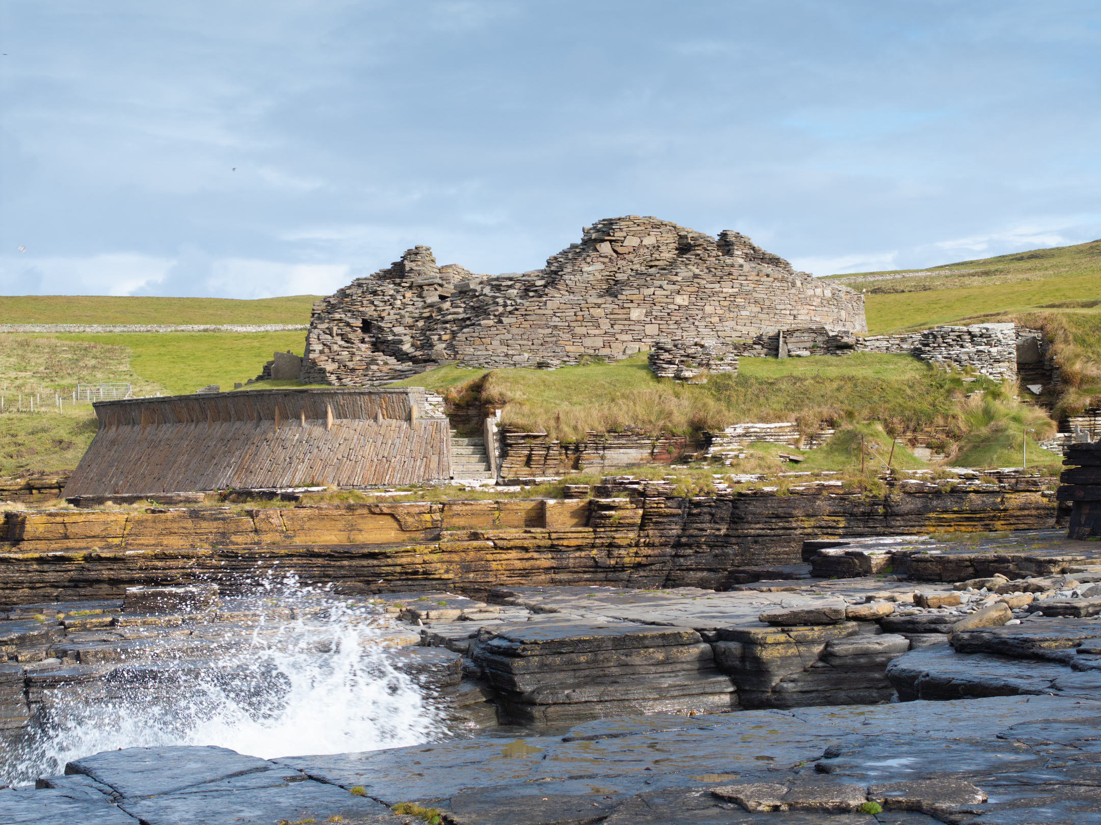 Midhowe Broch seen from the rocky shore, with waves splashing in the foreground and the broch’s crumbling stone structure rising behind.