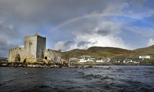 A castle sites on a small rocky island in the waters of Castlebay. The town and rolling hills are in the background, with a rainbow arching over a cloudy sky.