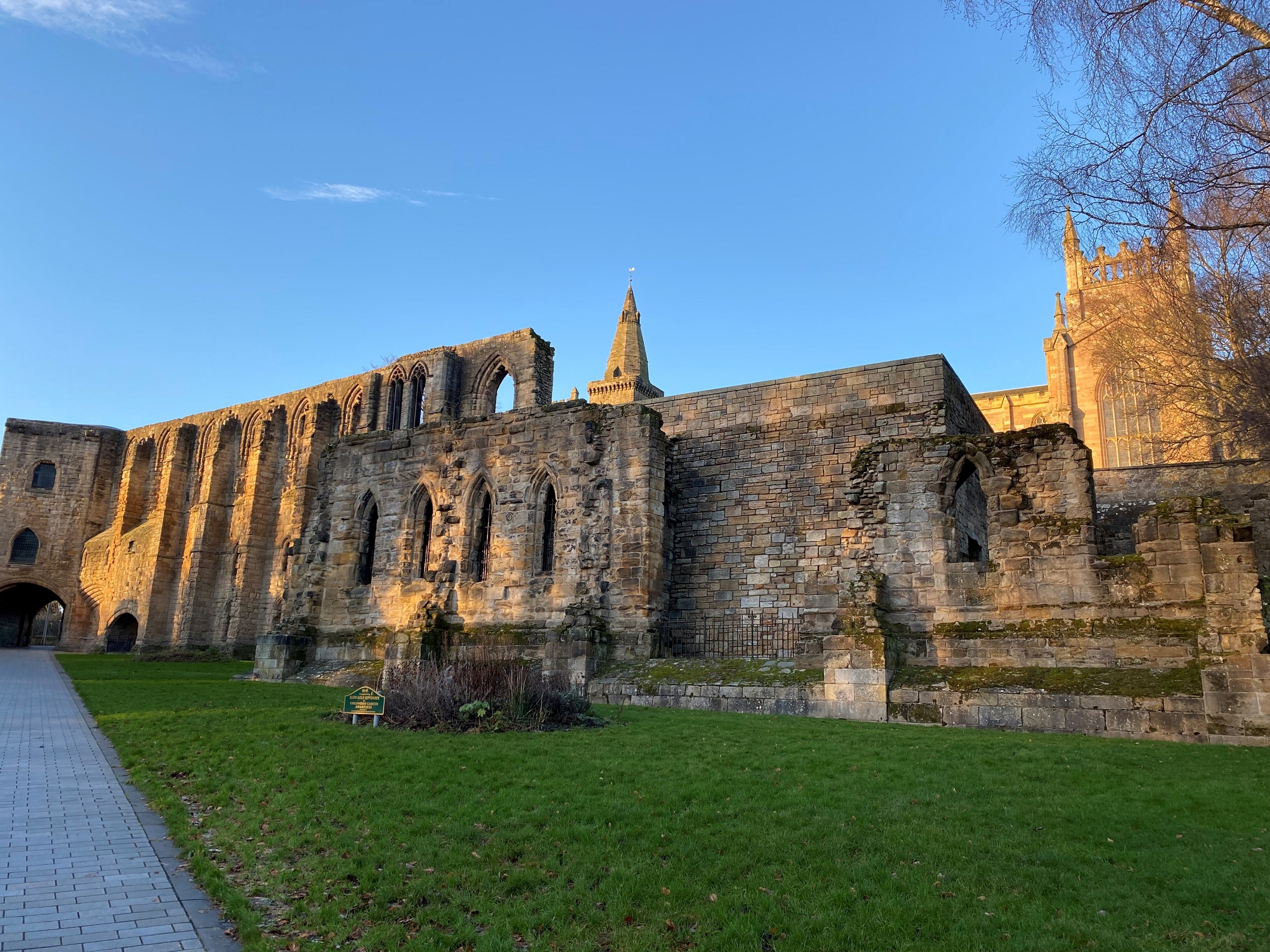 Warm evening light illuminates palace ruins with arched openings, set beside a paved path and grassy lawn.