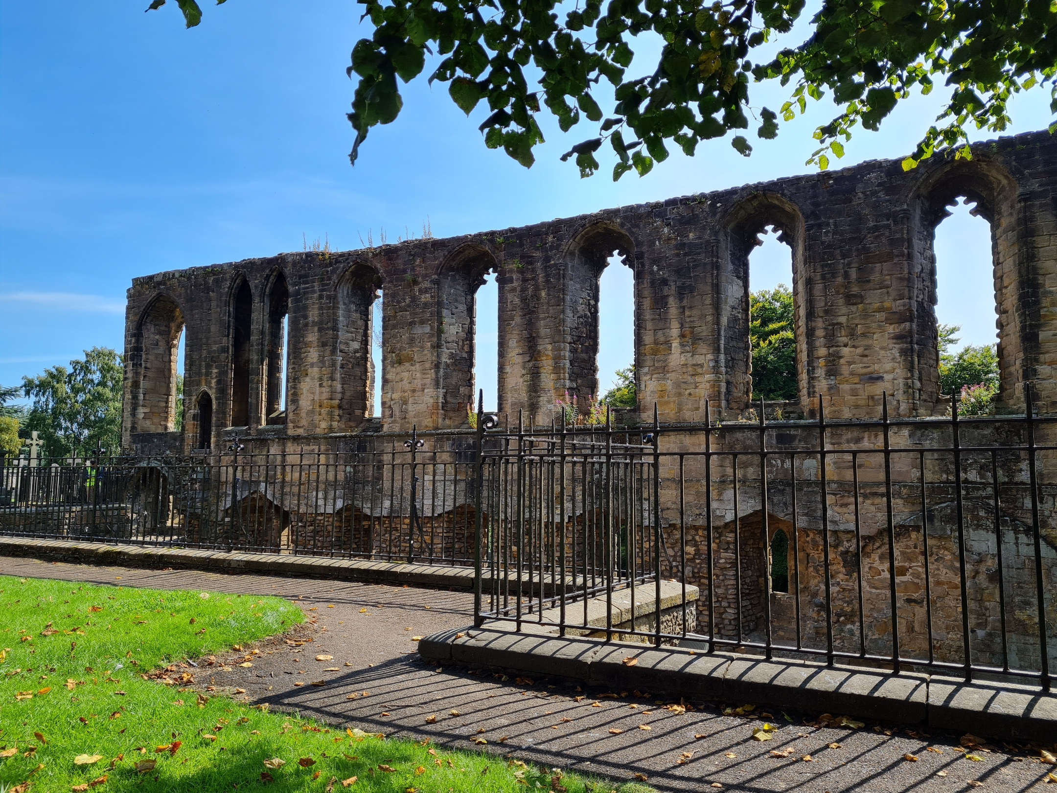 A long stretch of stone palace ruins with tall, narrow arched window openings, bordered by metal railings and surrounded by grass and trees.