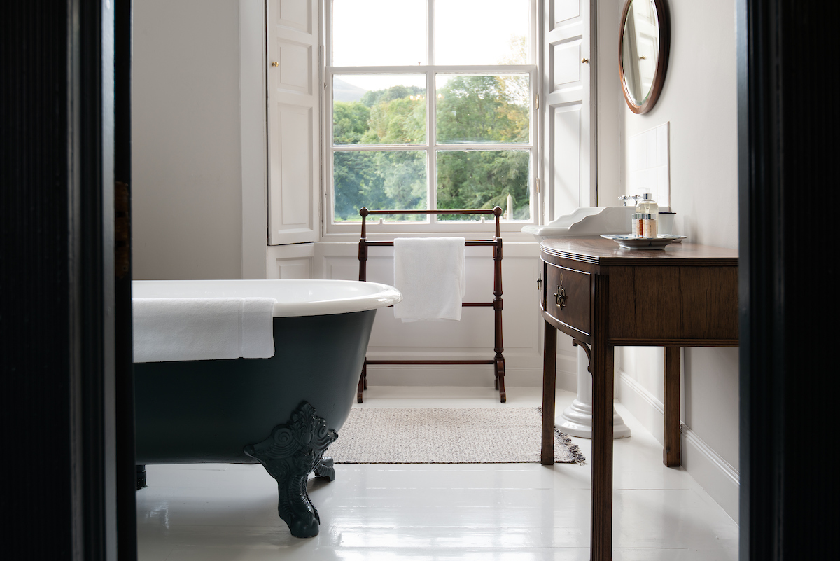 A white bathroom with a large, ironwork, black freestanding bath and large sash and case window looking out onto trees