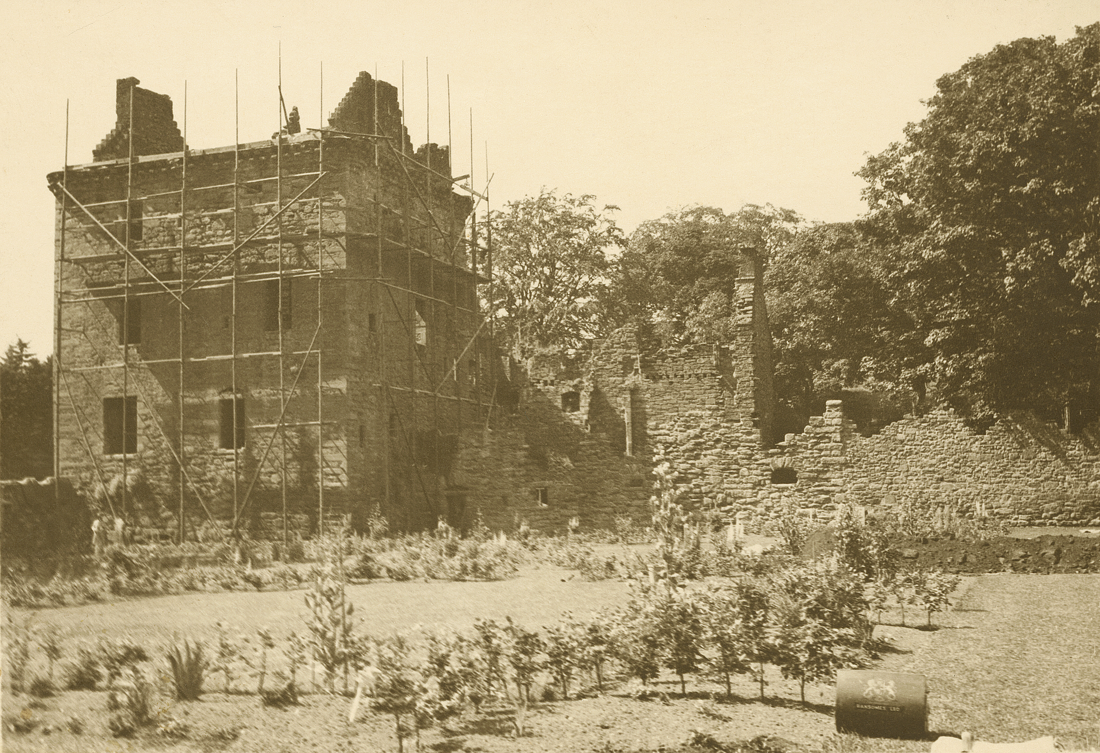 A black and white photo of a ruined castle and a walled garden, where hedges and shrubs have recently been planted. There is scaffolding on one side of the castle.