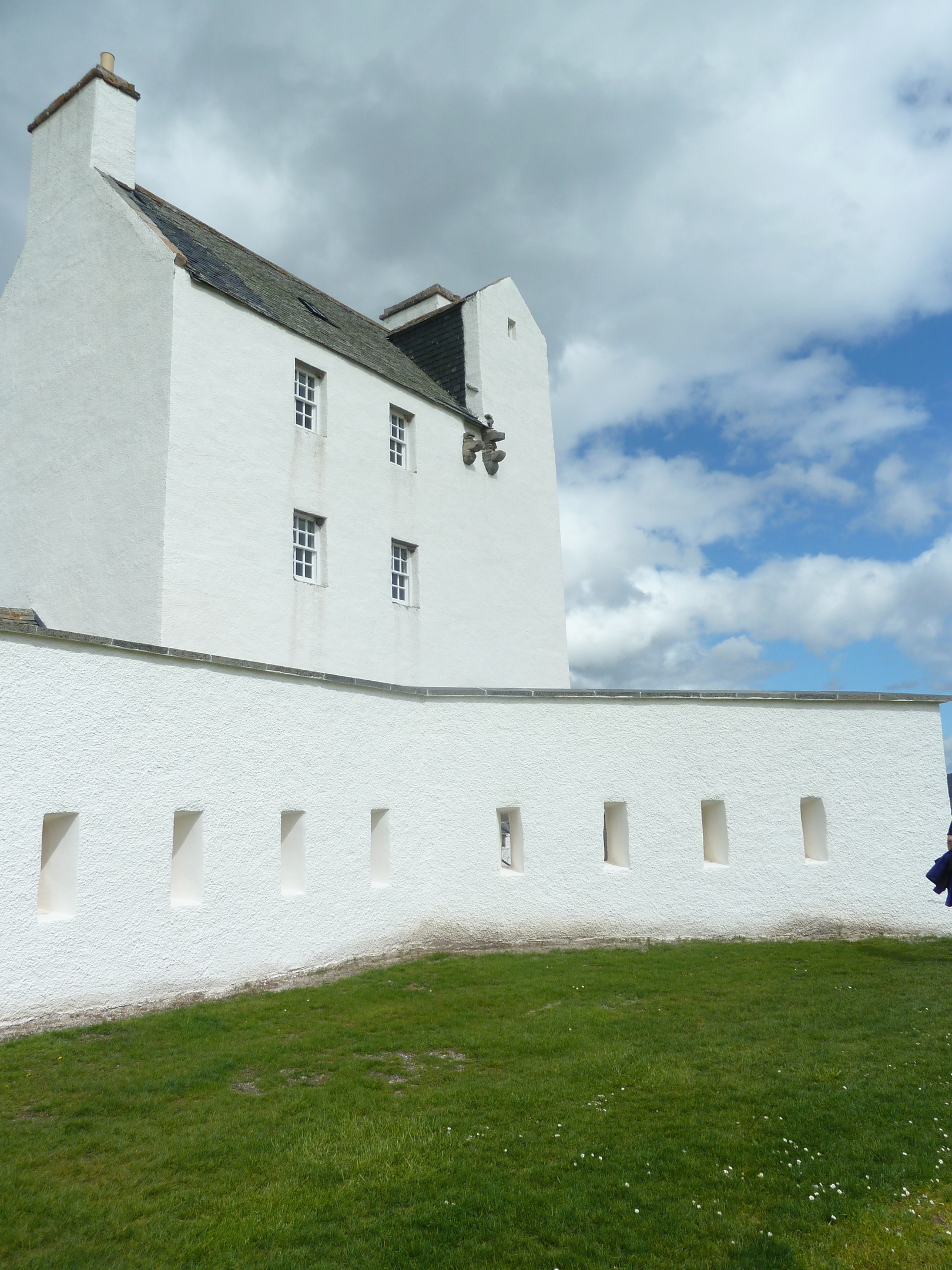 White exterior of Corgarff Castle’s tower and surrounding wall, viewed from the grassy outer area under a partly cloudy sky.