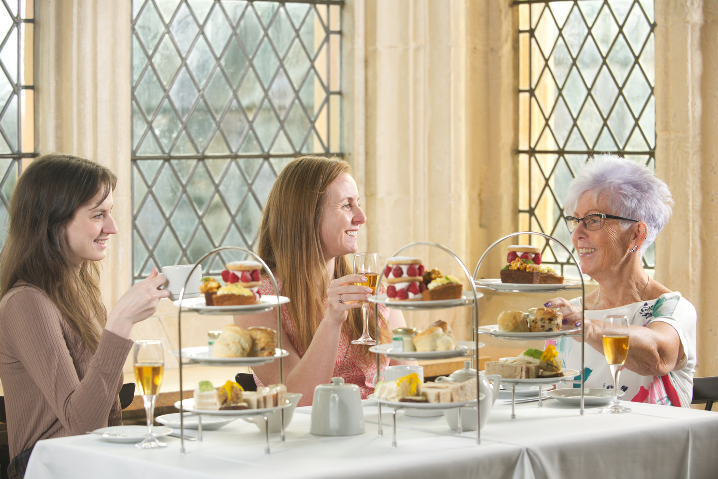 Three people smiling, sat at a table with afternoon tea in front of them. One person holds a mug, another holds a wine glass. There are cakes and sandwiches on stands. 